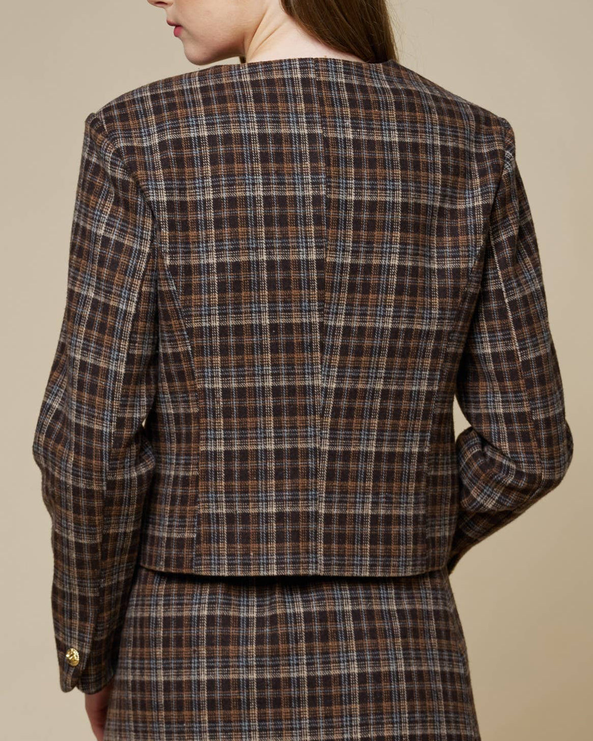 A woman shown from the back wearing a brown plaid blazer and matching mini skirt against a solid beige studio background.