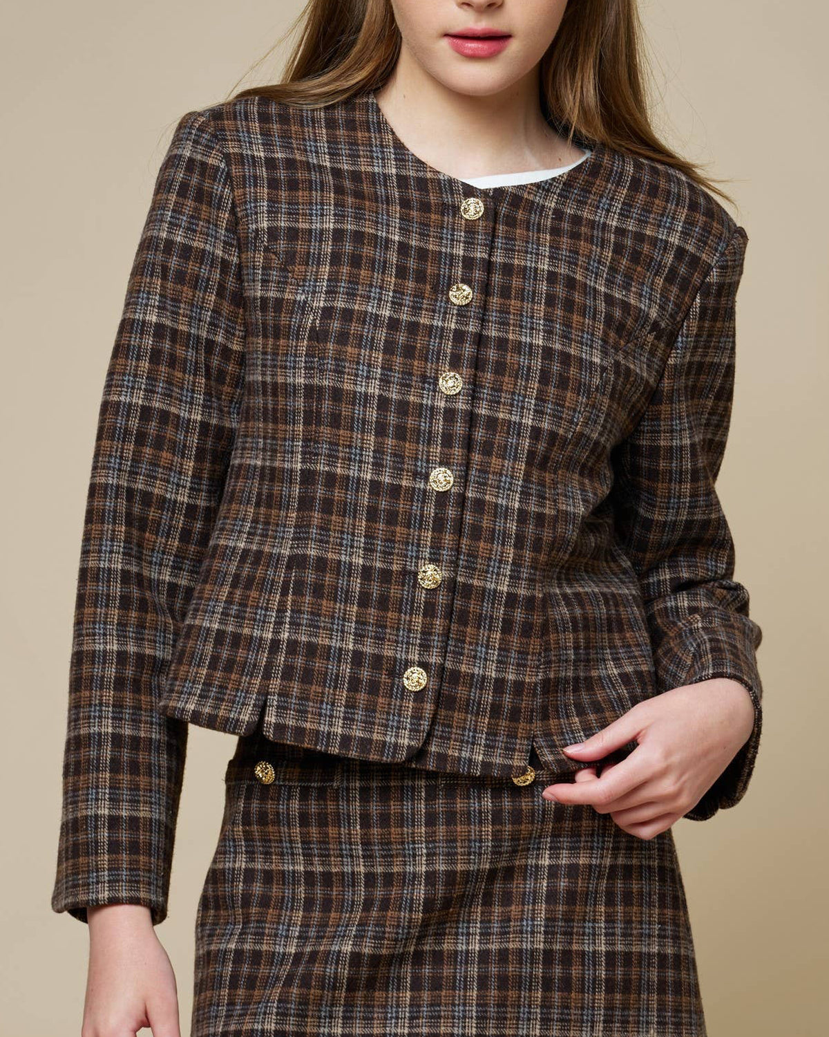 A young woman modeling a matching brown plaid tweed jacket and mini skirt set against a solid beige studio background.