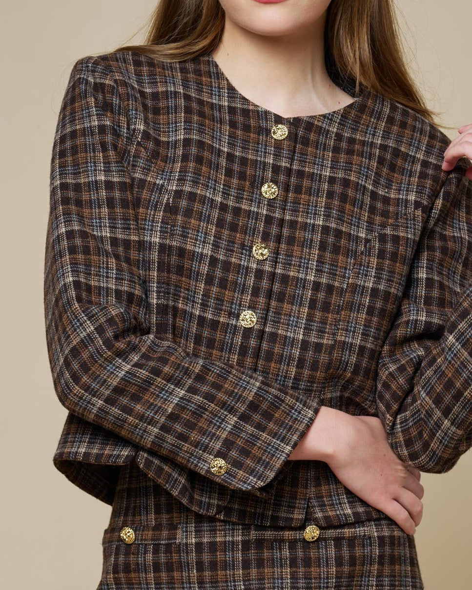 A young woman modeling a brown and beige plaid mini dress with gold buttons, posed against a solid beige studio background.