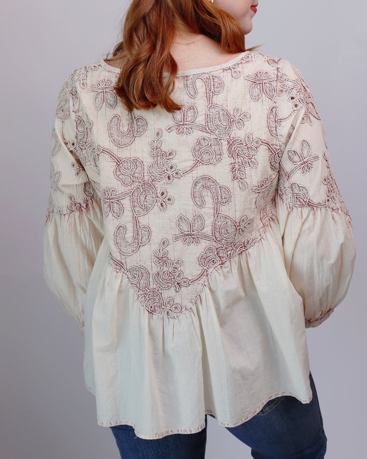 A woman wearing a cream-colored long-sleeve blouse with intricate dusty rose embroidery on the back, paired with blue jeans, shown from a rear three-quarter view against a plain gray background.