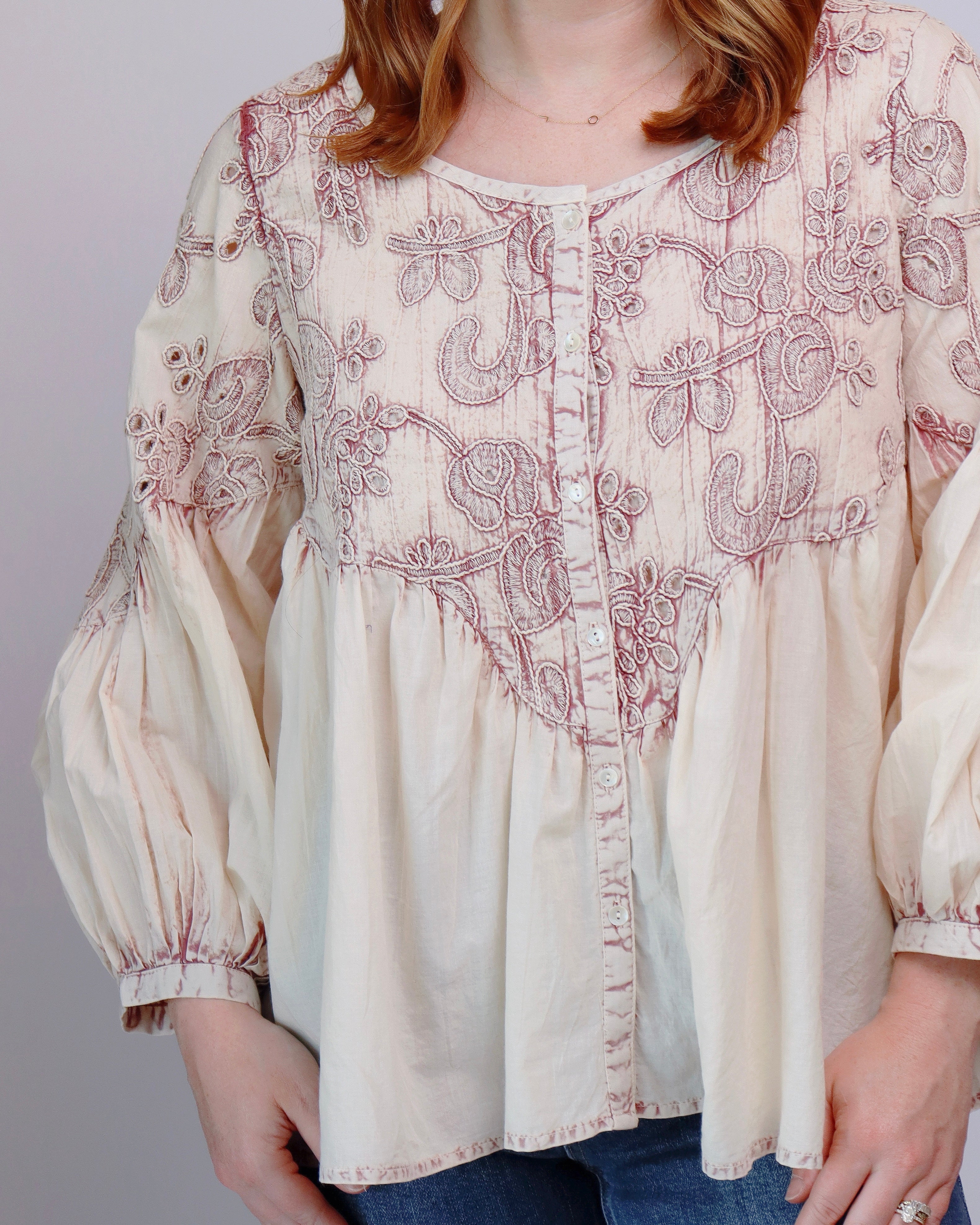 A woman wearing a cream-colored long-sleeve blouse with dusty rose embroidery and a button-down front, paired with blue jeans.