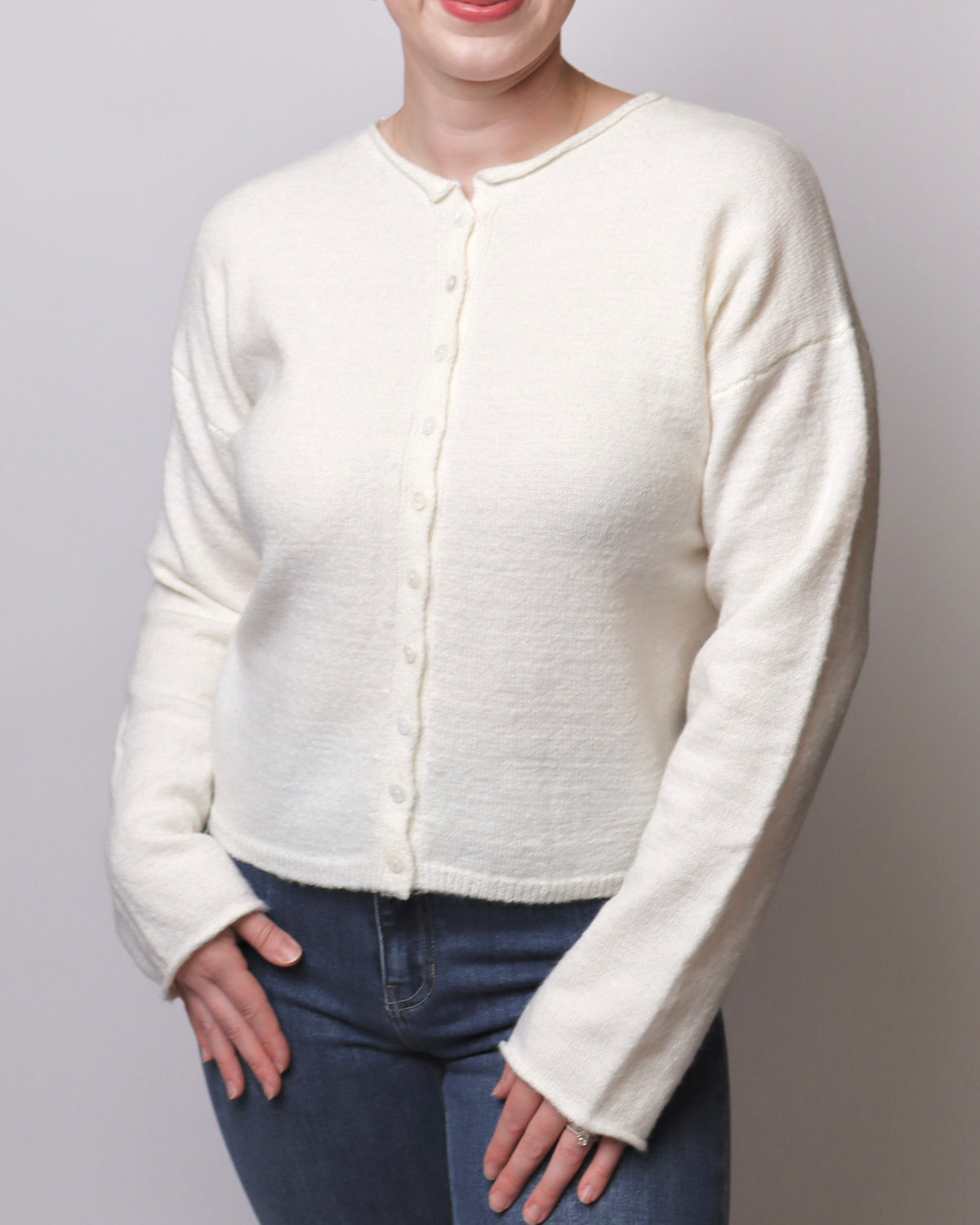 A woman wearing a cream-colored, long-sleeve button-up knit cardigan sweater and dark blue jeans, shown from the chin down against a neutral light gray background.