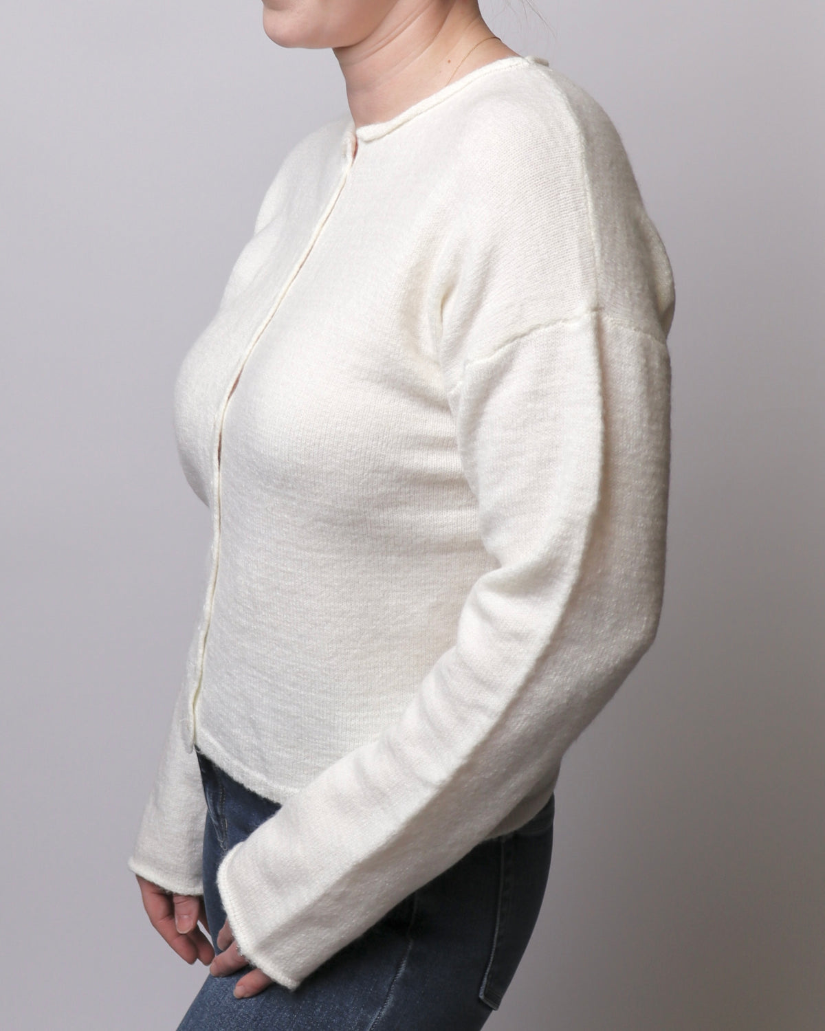 A woman wearing a cream-colored, long-sleeve knit cardigan with a round neckline and front buttons, paired with dark blue jeans, shown from a side profile against a neutral gray background.