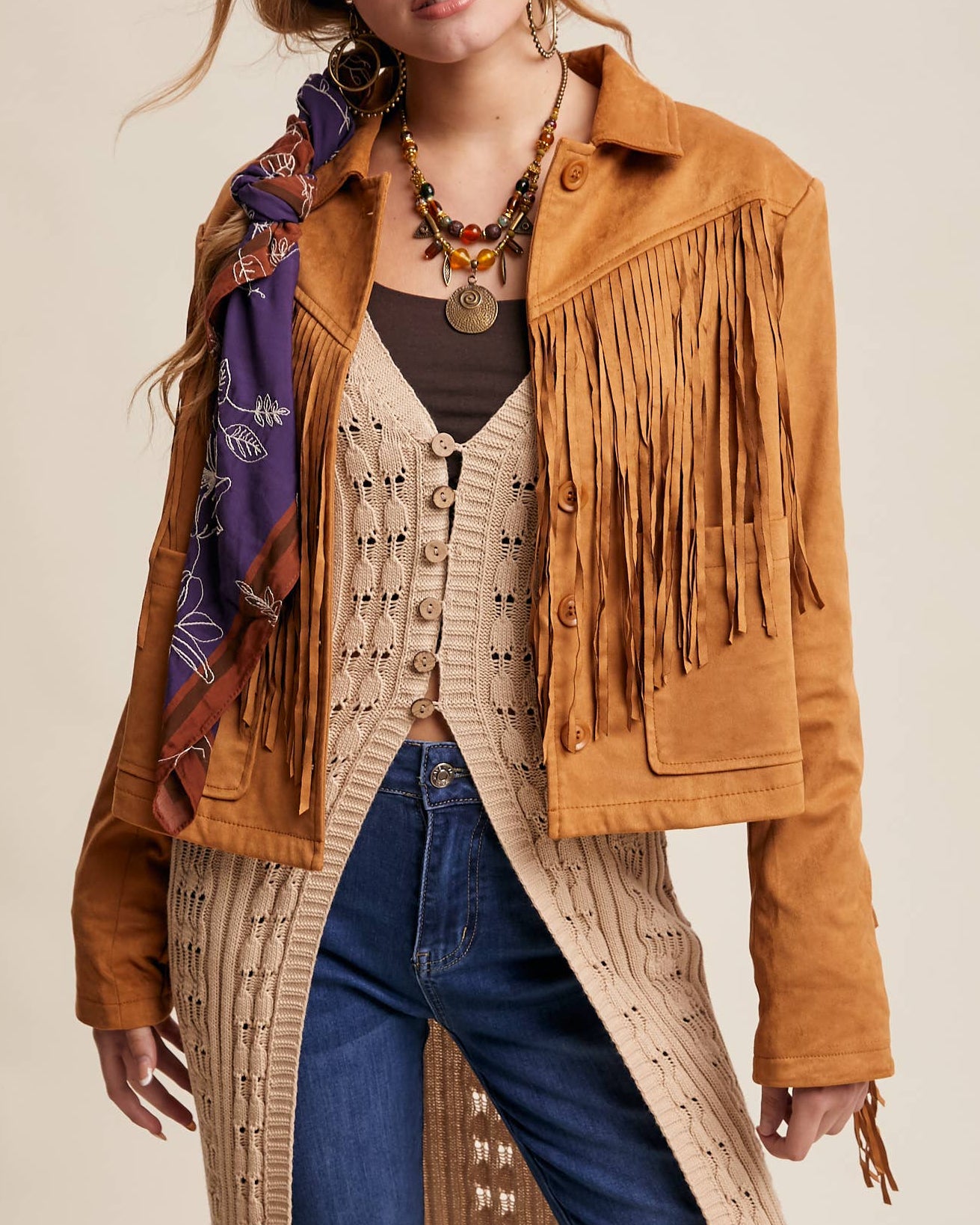 Woman wearing a brown fringed jacket, blue jeans, and a patterned dress with a plain background