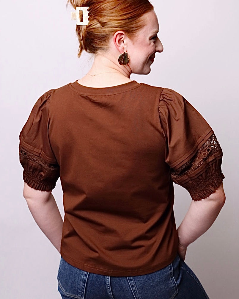 A woman shown from the back wearing a brown short-sleeve top with textured lace trim at the sleeve cuffs, paired with blue jeans, against a plain light gray background.