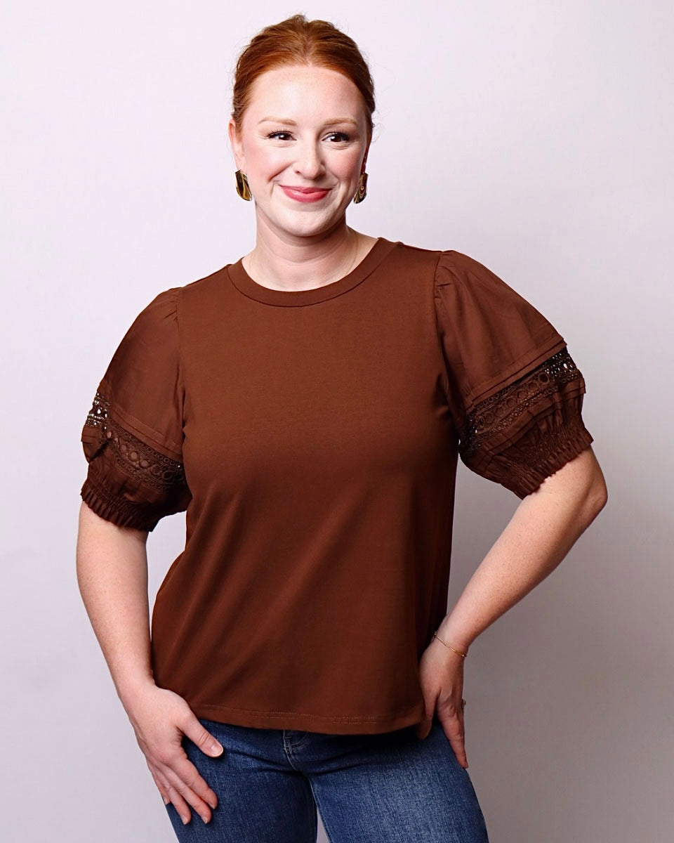 A woman modeling a brown short-sleeve top with sheer puff sleeves and lace detailing on the cuffs, paired with blue jeans against a light gray studio background.