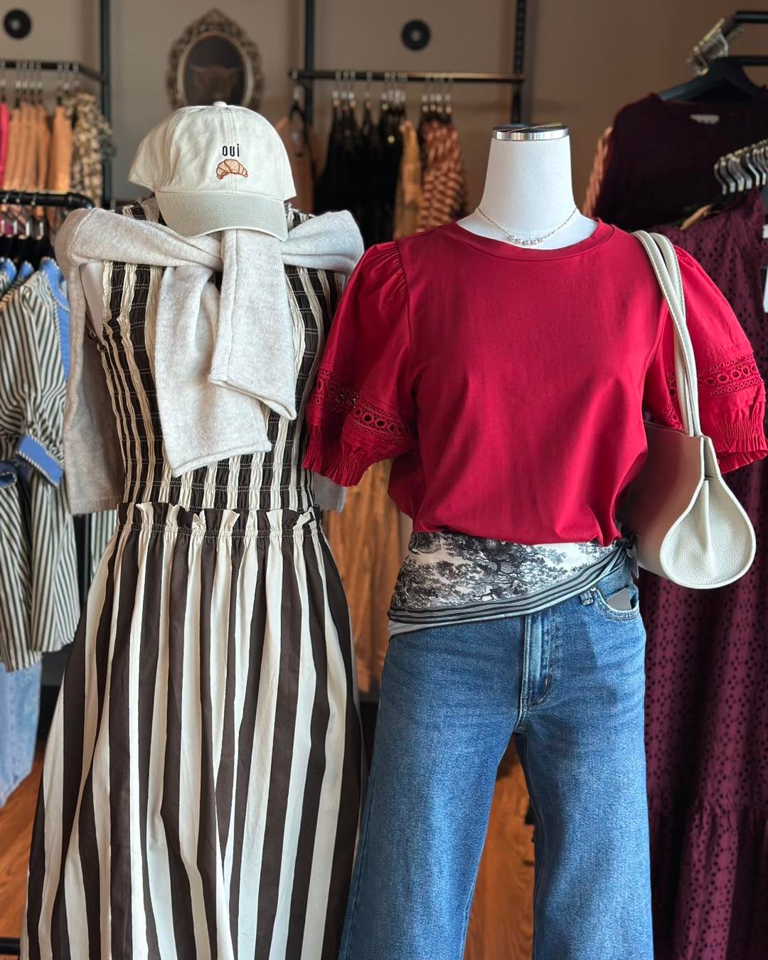 A mannequin in a clothing boutique wearing a bright red puff-sleeve top with eyelet lace details, blue jeans, a patterned waist layer, and a cream shoulder bag. Another mannequin wears a brown and white striped dress with a cream cardigan and cap.