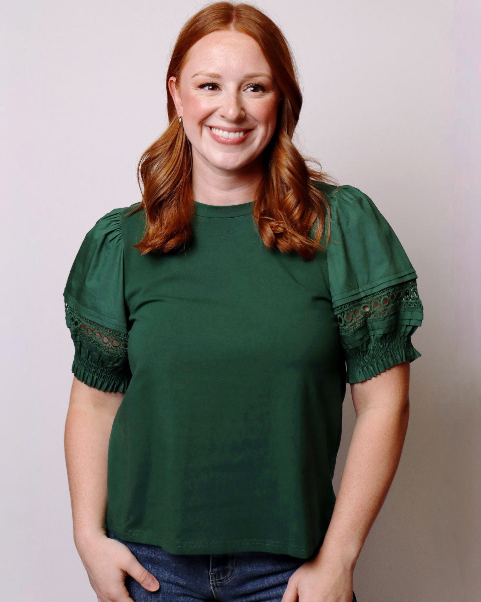 A smiling woman with red hair wearing a dark green short-sleeve top with lace-up puff sleeves and blue jeans, standing against a plain light gray background.