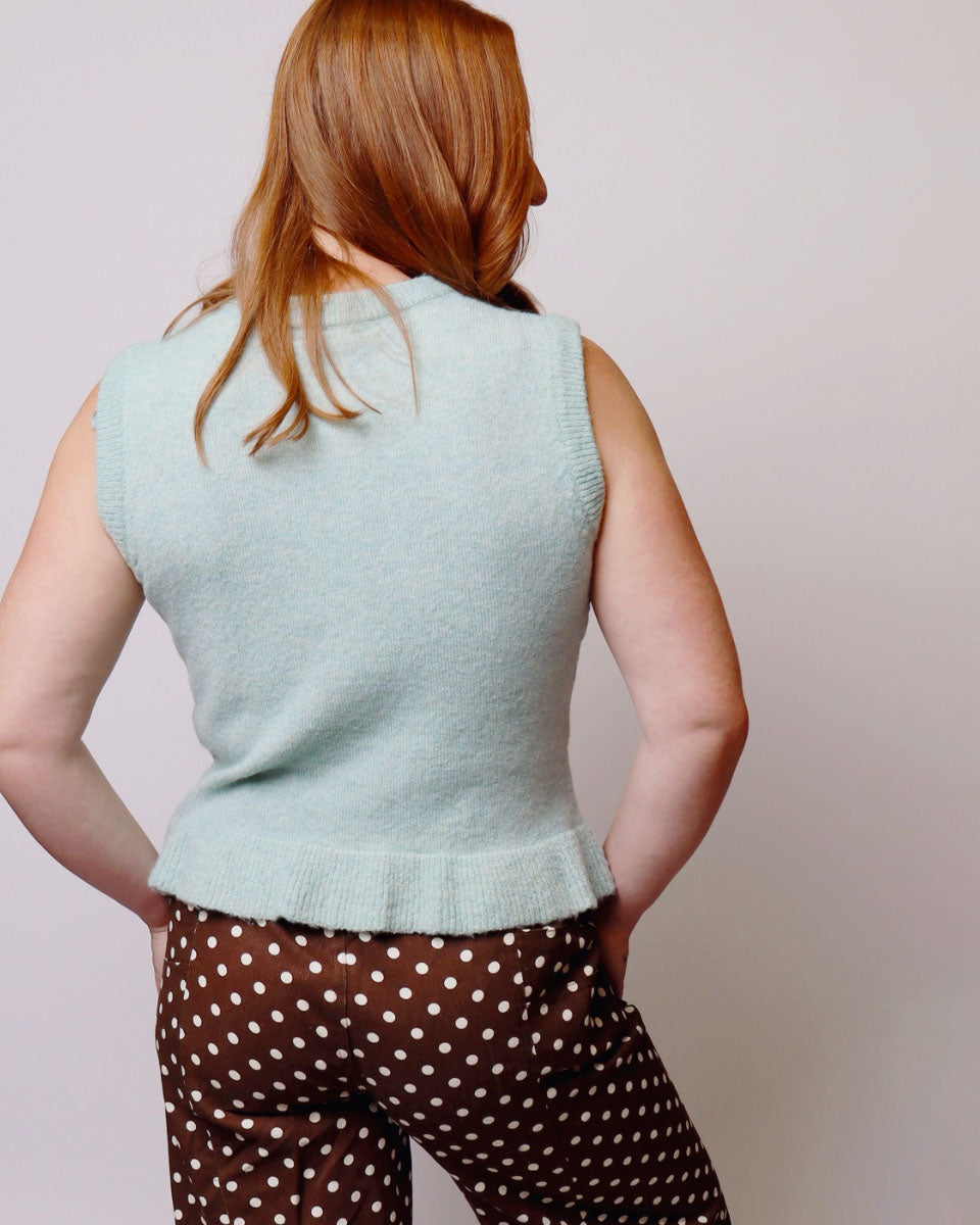 A woman shown from behind wearing a light sage green sleeveless knit vest and brown pants with white polka dots, against a plain light gray background.