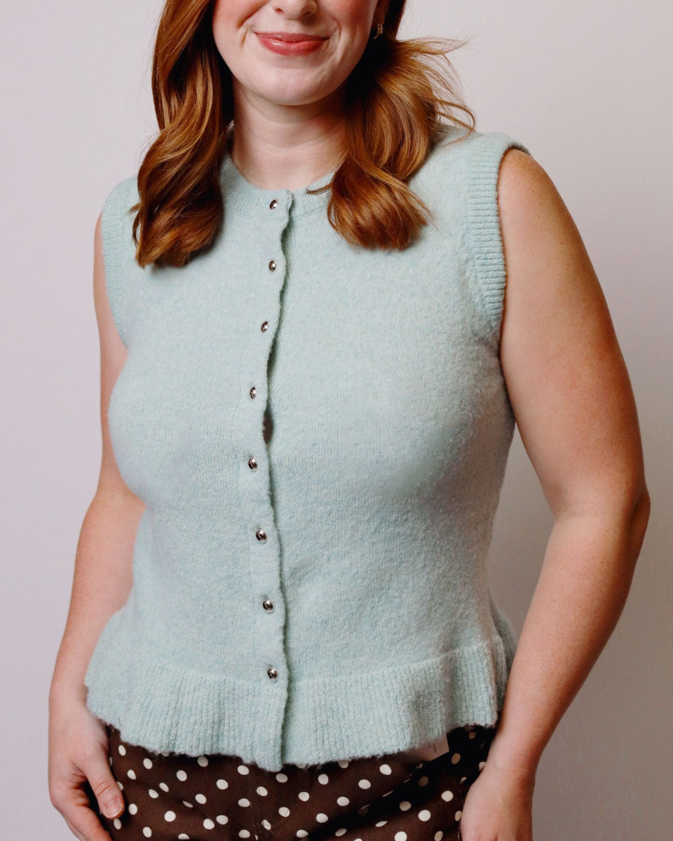 A woman wearing a sleeveless, ribbed knit cardigan vest in a pale mint green color, paired with brown and white polka-dot pants, against a plain light gray background.