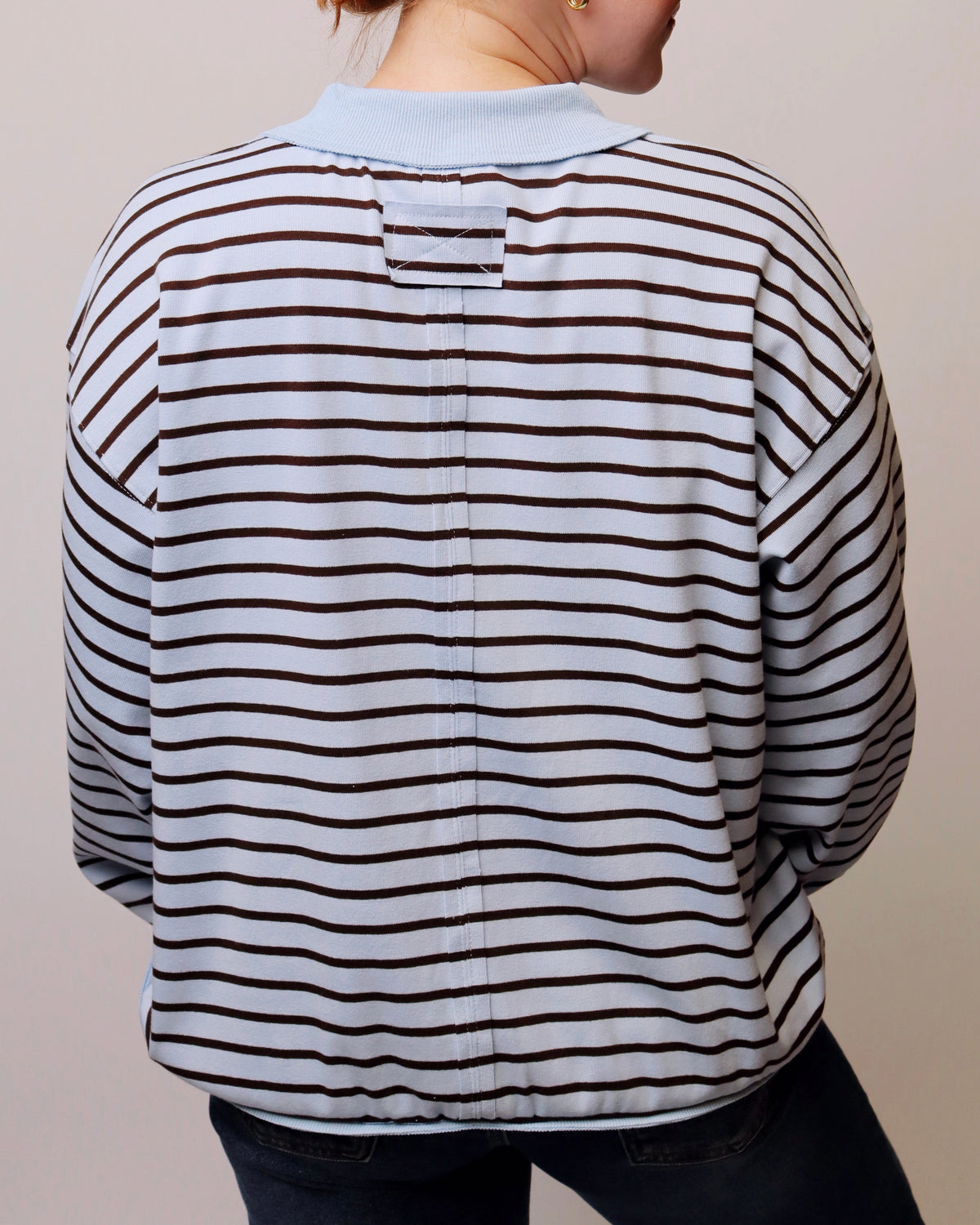 A person shown from the back wearing a long-sleeve collared shirt with thin dark brown horizontal stripes on a white base, paired with dark jeans against a plain light beige background.