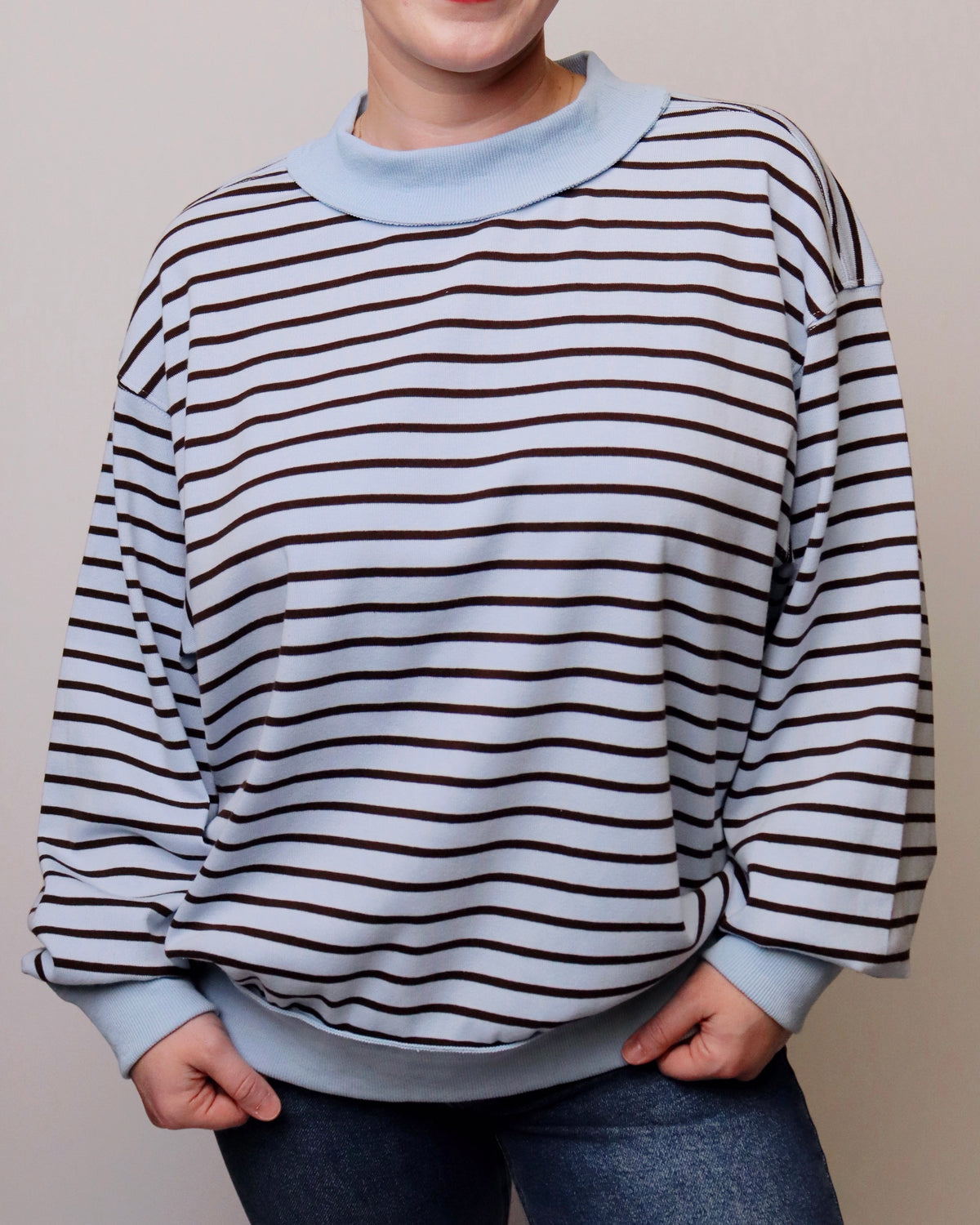 A person wearing a light blue crewneck sweatshirt with thin dark brown horizontal stripes, paired with blue jeans, shown from the neck down against a plain beige background.