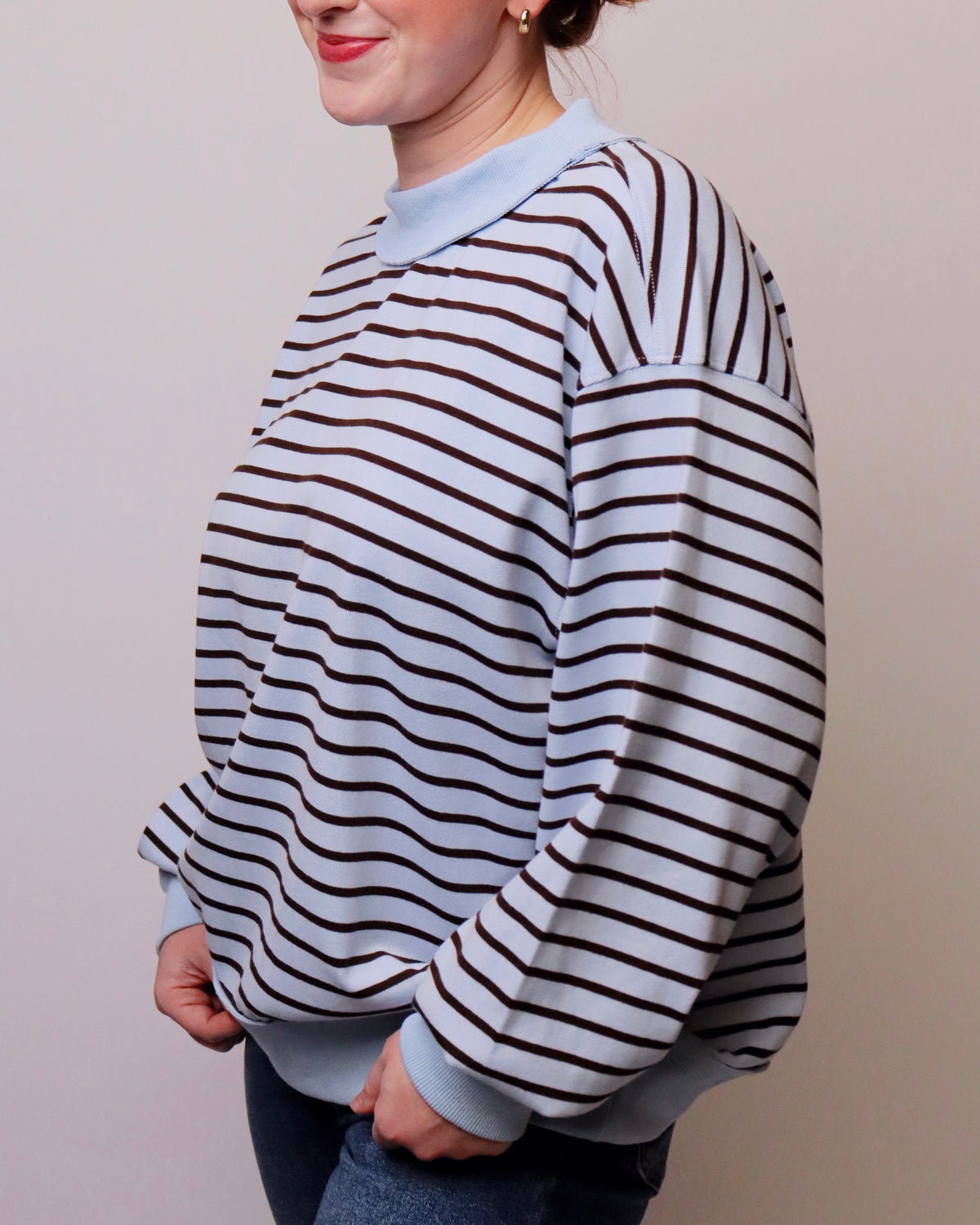 A person wearing a long-sleeve crewneck sweatshirt with horizontal dark brown and white stripes, paired with a light blue ribbed collar, cuffs, and hem, shown against a plain light beige background.