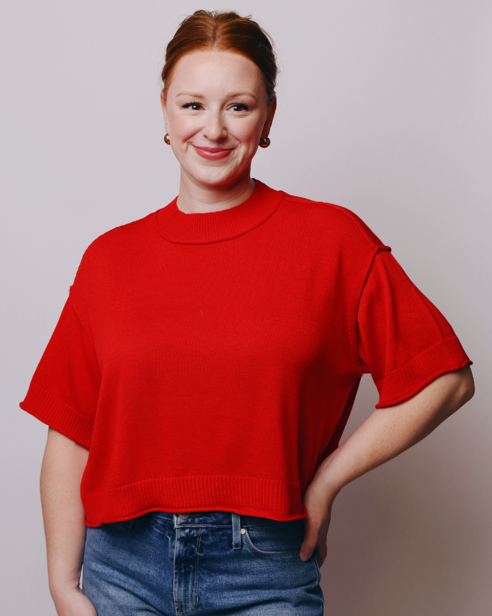 A woman with red hair tied back, wearing a bright red short-sleeve knit sweater and blue jeans, posing against a light gray studio background.