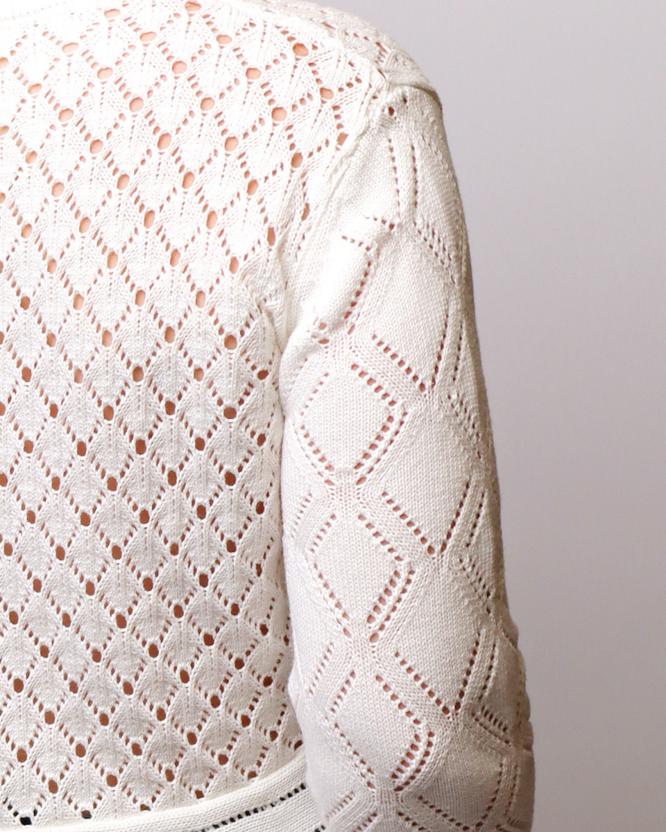 Close-up of a cream-colored knit sweater featuring a textured geometric diamond pattern with openwork details on the body and sleeves.