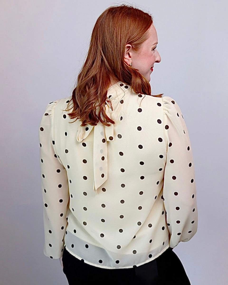 A woman with red hair is shown from the back wearing a cream-colored long-sleeve blouse with black polka dots and a bow detail at the back neckline, paired with black pants against a plain light gray background.