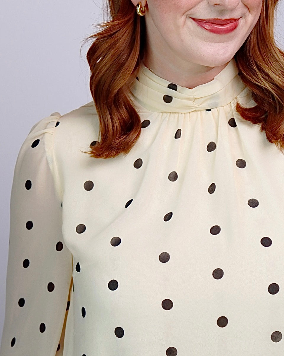 A close-up of a woman wearing a cream-colored long-sleeve blouse with black polka dots and a high gathered mock neck.