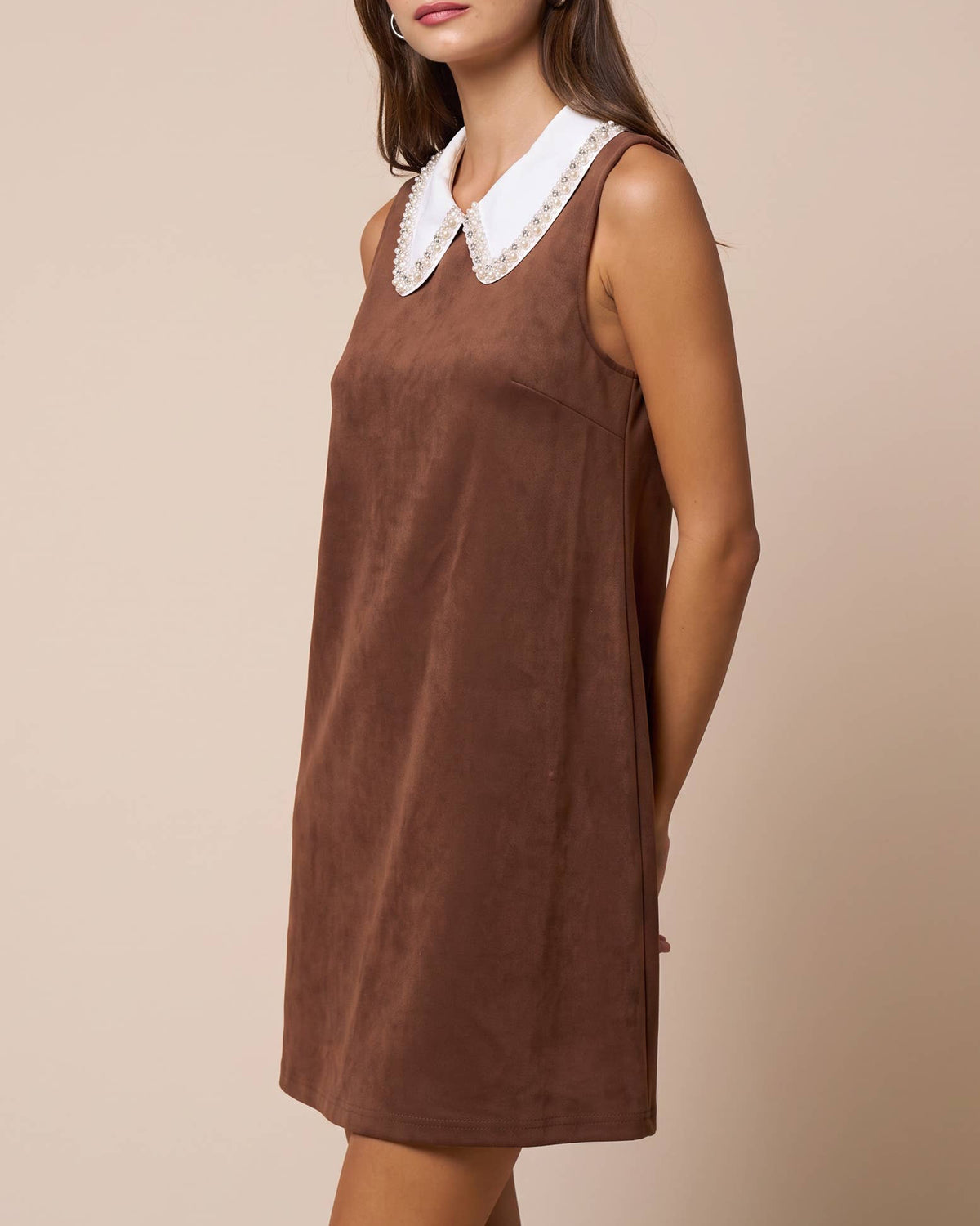 A woman modeling a sleeveless brown dress with a white lace Peter Pan collar, posed against a solid beige studio background. Isabella Rhinestone Contrast Collar Faux Suede Mini Shift Dress | Blackbird Boutique