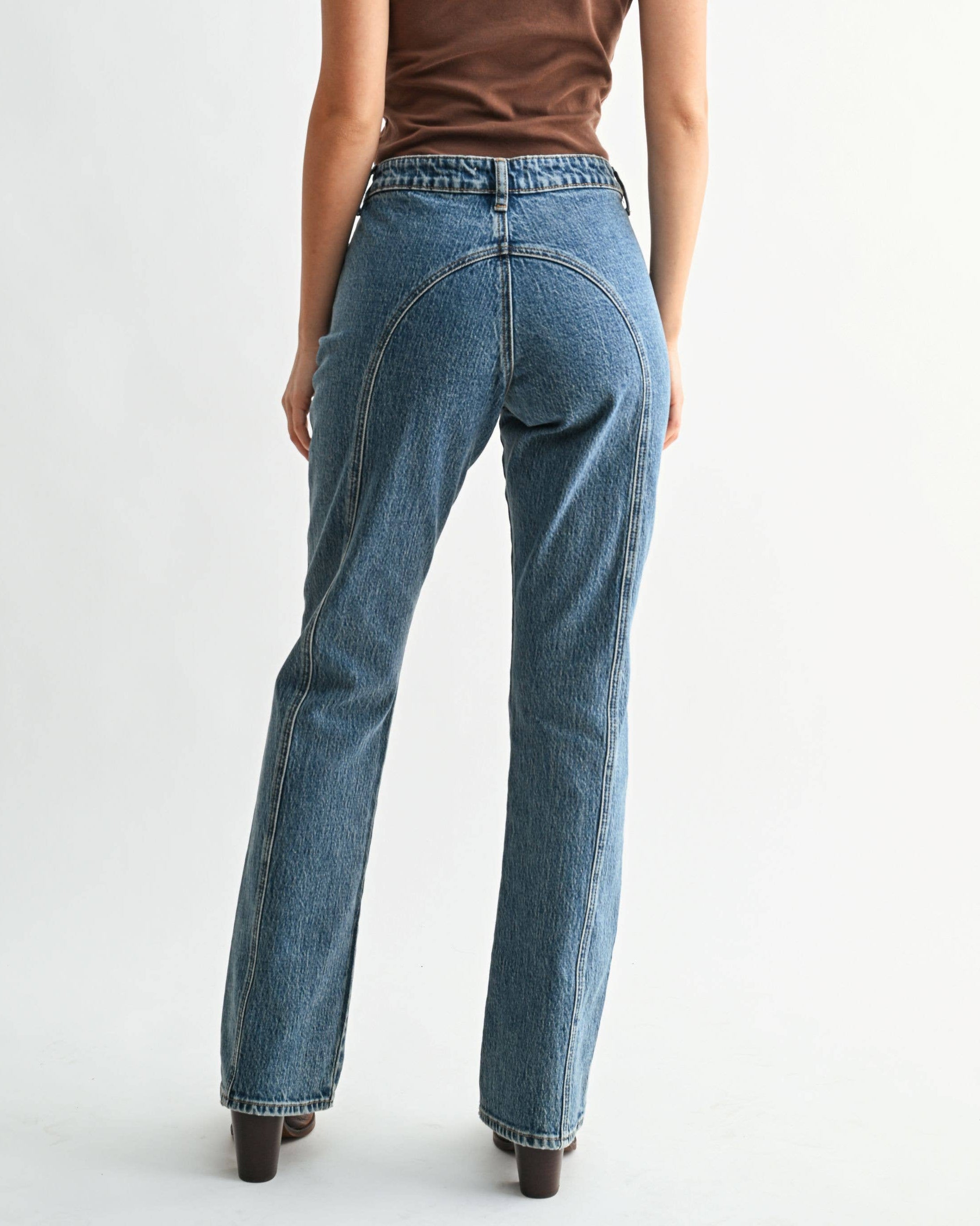 A woman shown from the back wearing a brown short-sleeve t-shirt and blue straight-leg jeans, standing against a plain light gray studio background.
