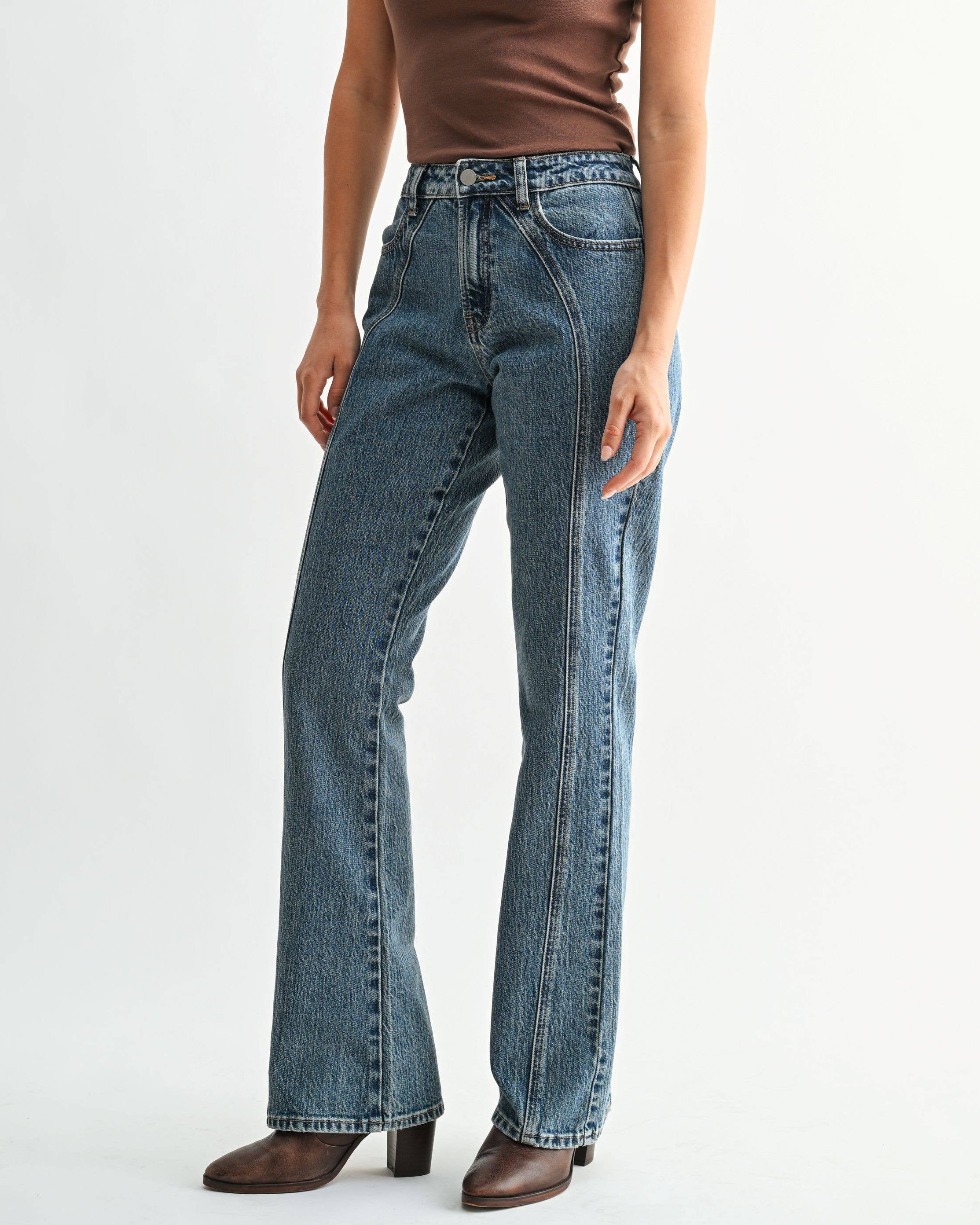 A woman modeling a brown short-sleeve t-shirt with a small white graphic on the chest, paired with high-waisted blue flare jeans and brown heeled boots, against a plain white studio background.