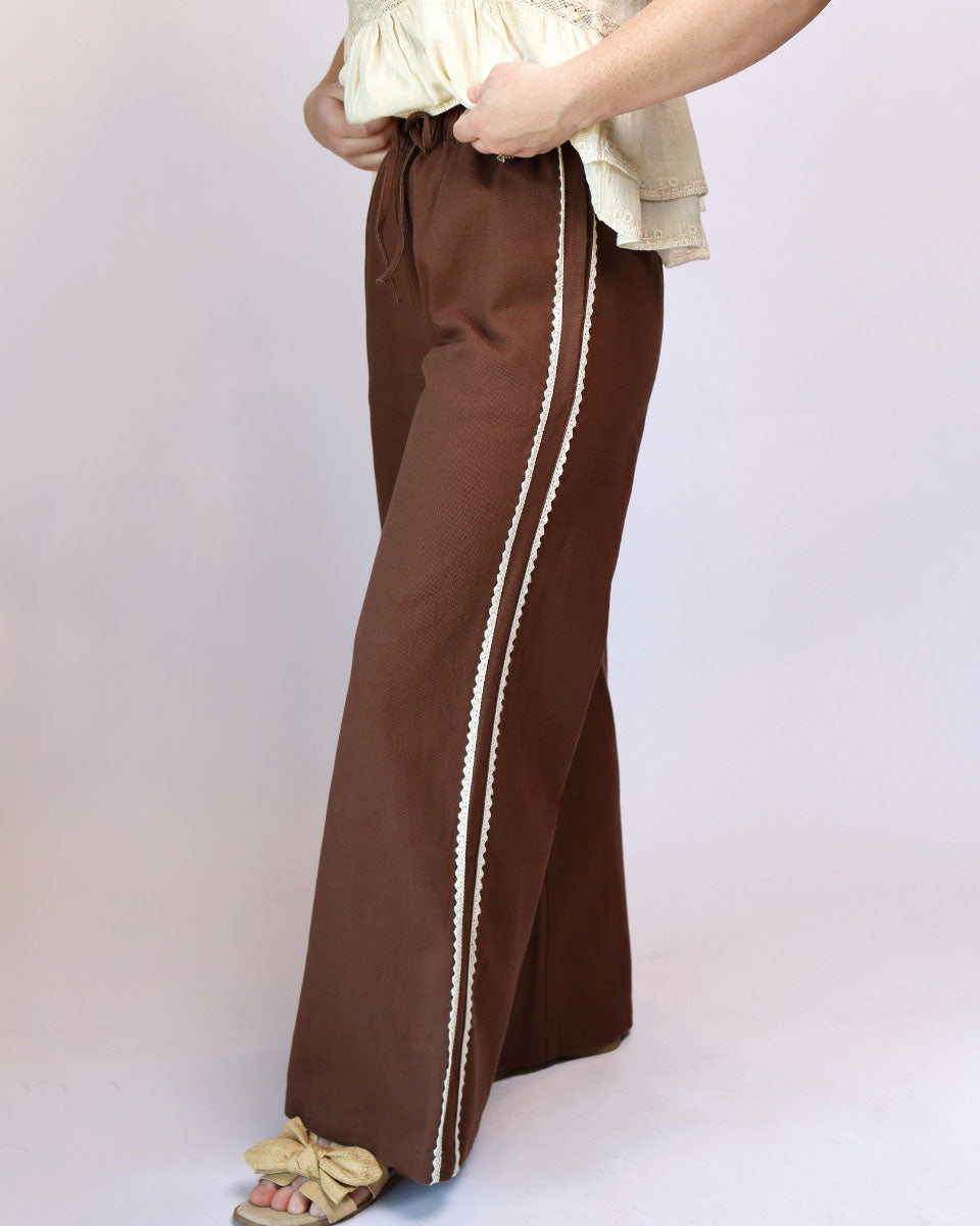 A person wearing brown wide-leg pants with a cream-colored braided stripe down the side, paired with a cream top and beige sandals, against a light studio background.