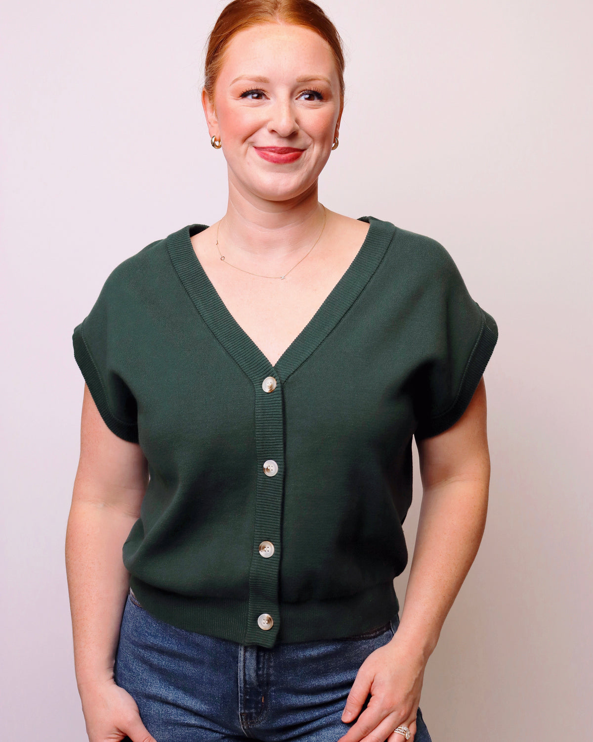 A woman with red hair tied back, wearing a dark green short-sleeve button-up knit top and blue jeans, standing against a light gray studio background.