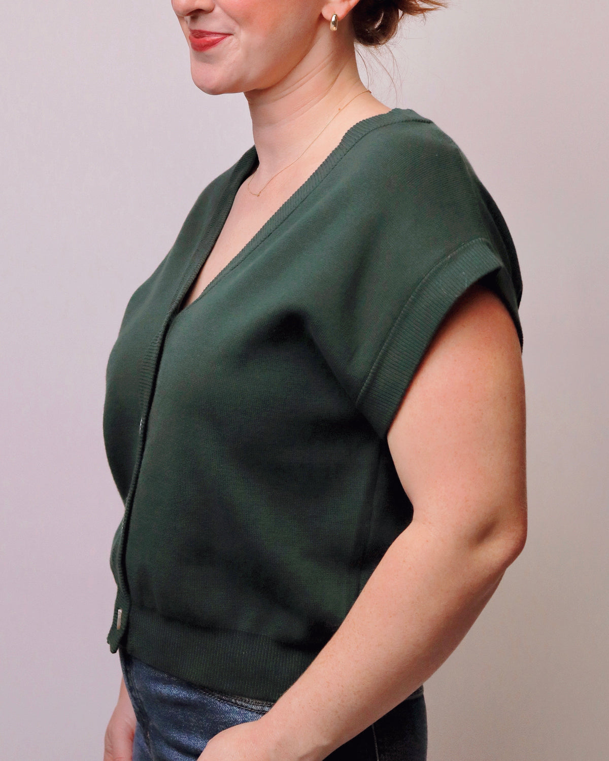 A woman wearing a dark green knit cardigan with short sleeves and a front zipper, shown from the side against a plain light background.