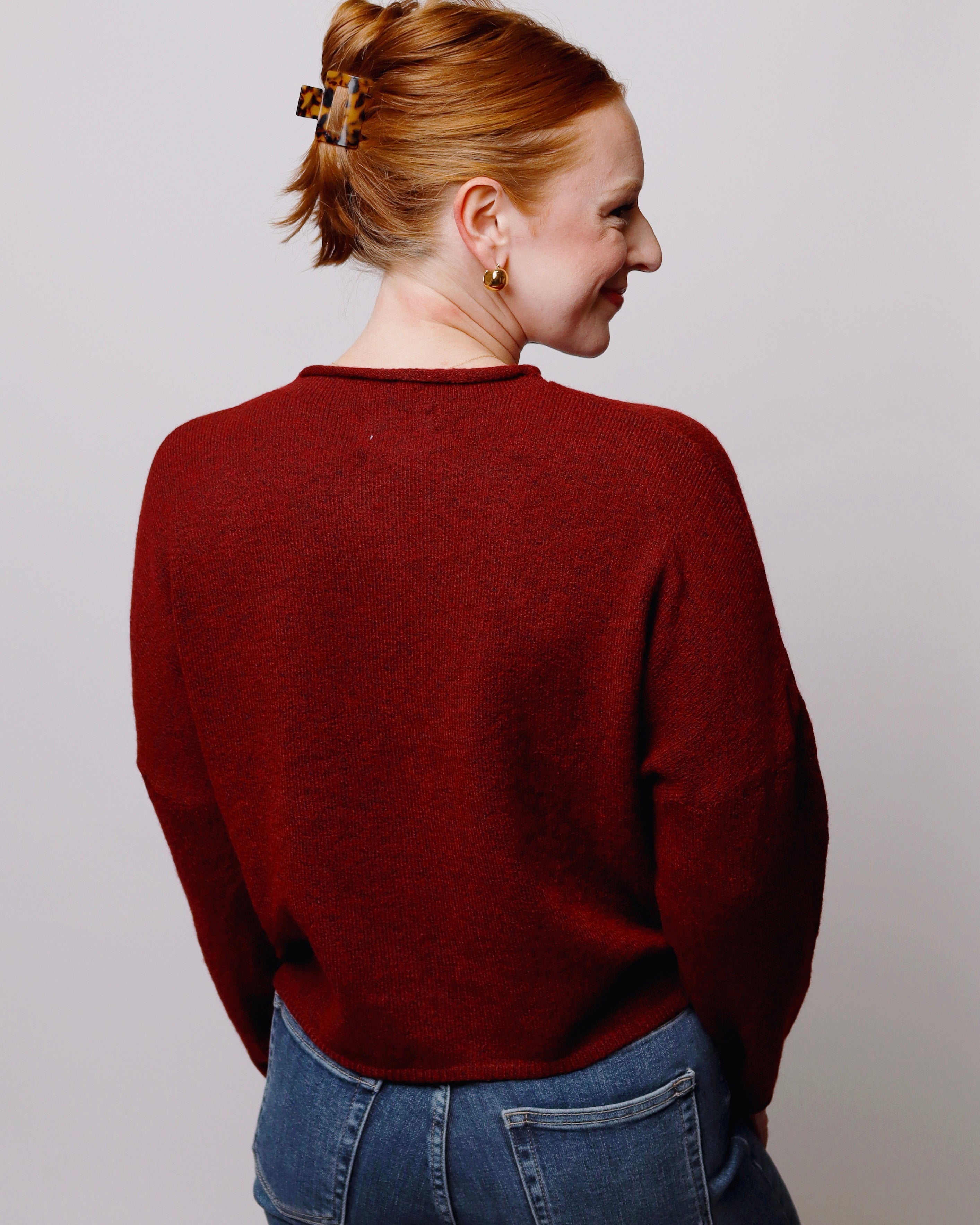 A woman with red hair tied back in a bun is shown from the back wearing a dark red knit sweater and blue jeans against a plain light gray background.