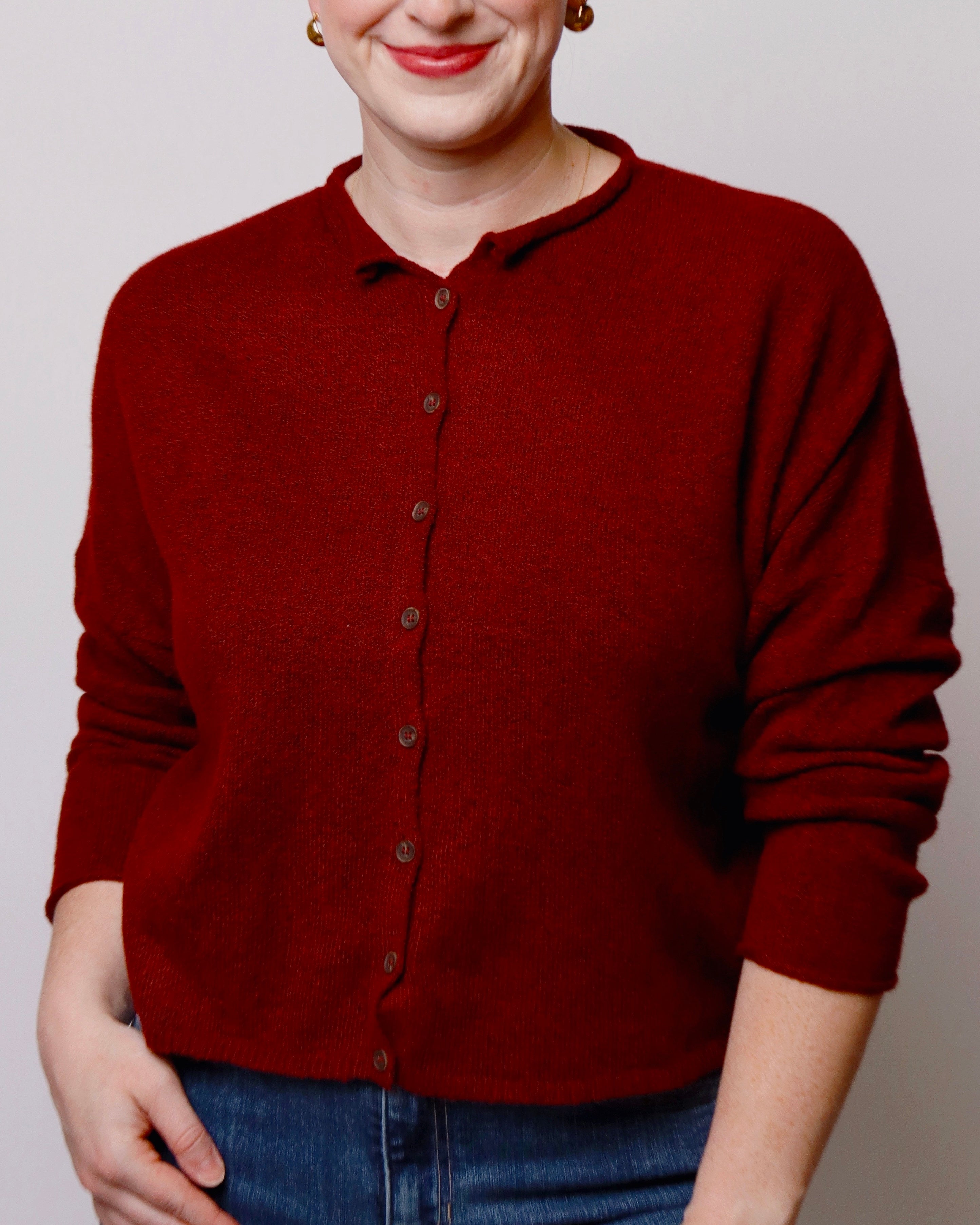 A woman wearing a dark red button-up cardigan sweater with a relaxed fit and blue jeans, shown from the nose down against a plain light gray background.
