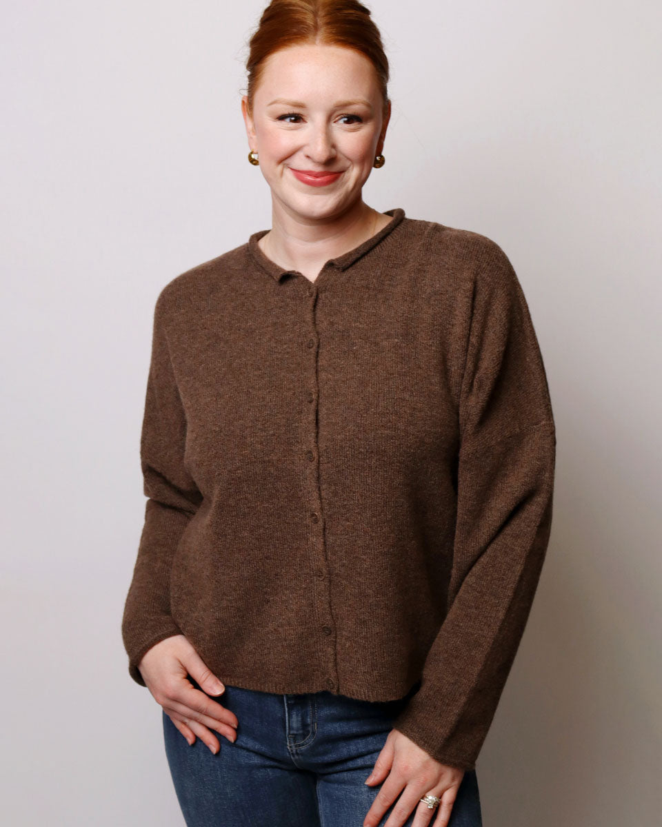 A woman with red hair wearing a brown button-up knit cardigan sweater and blue jeans, standing against a light gray studio background.