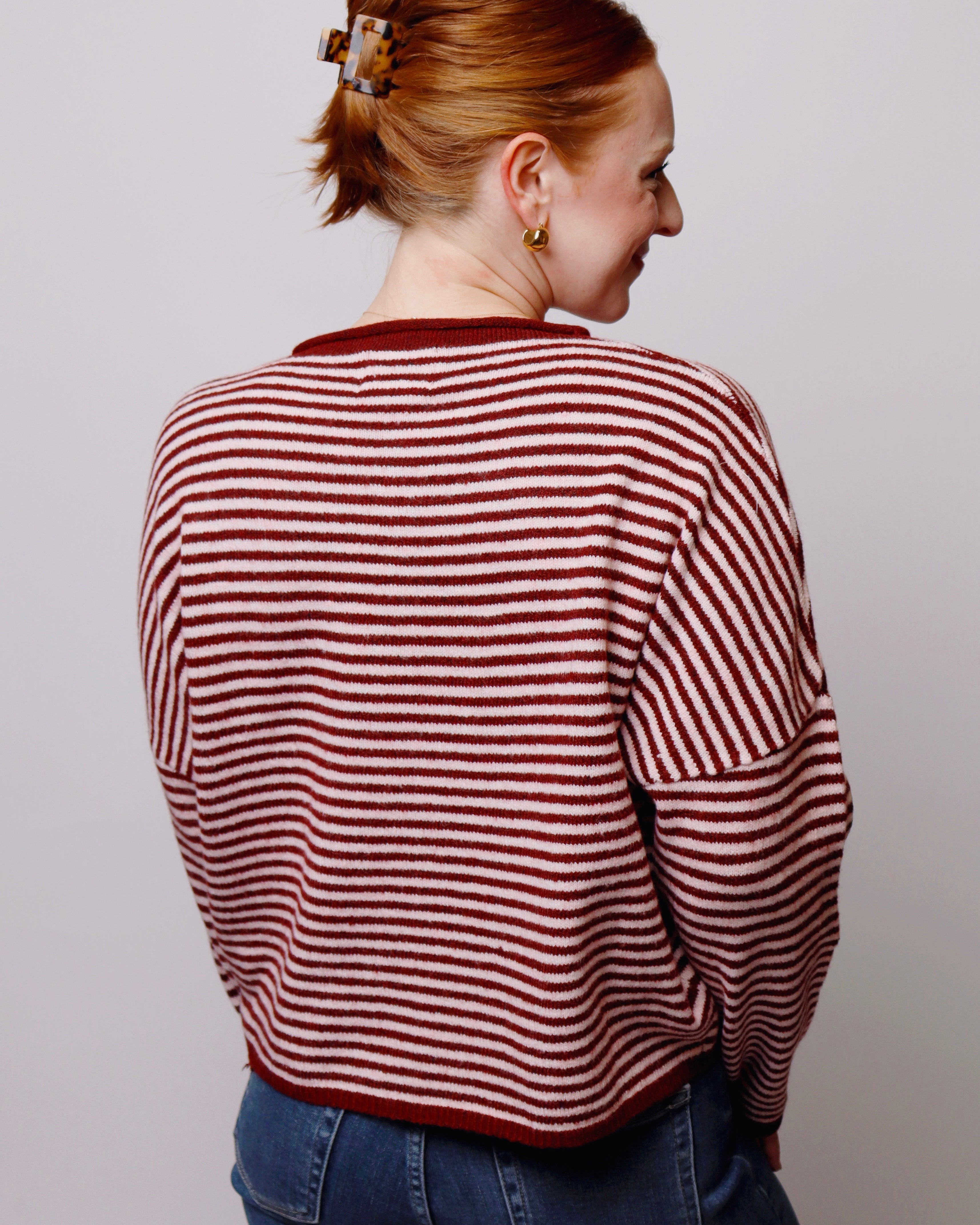 A woman with red hair tied up in a bun is shown from the back wearing a cropped, long-sleeve knit sweater with horizontal red and pink stripes, paired with blue jeans against a plain light gray background.