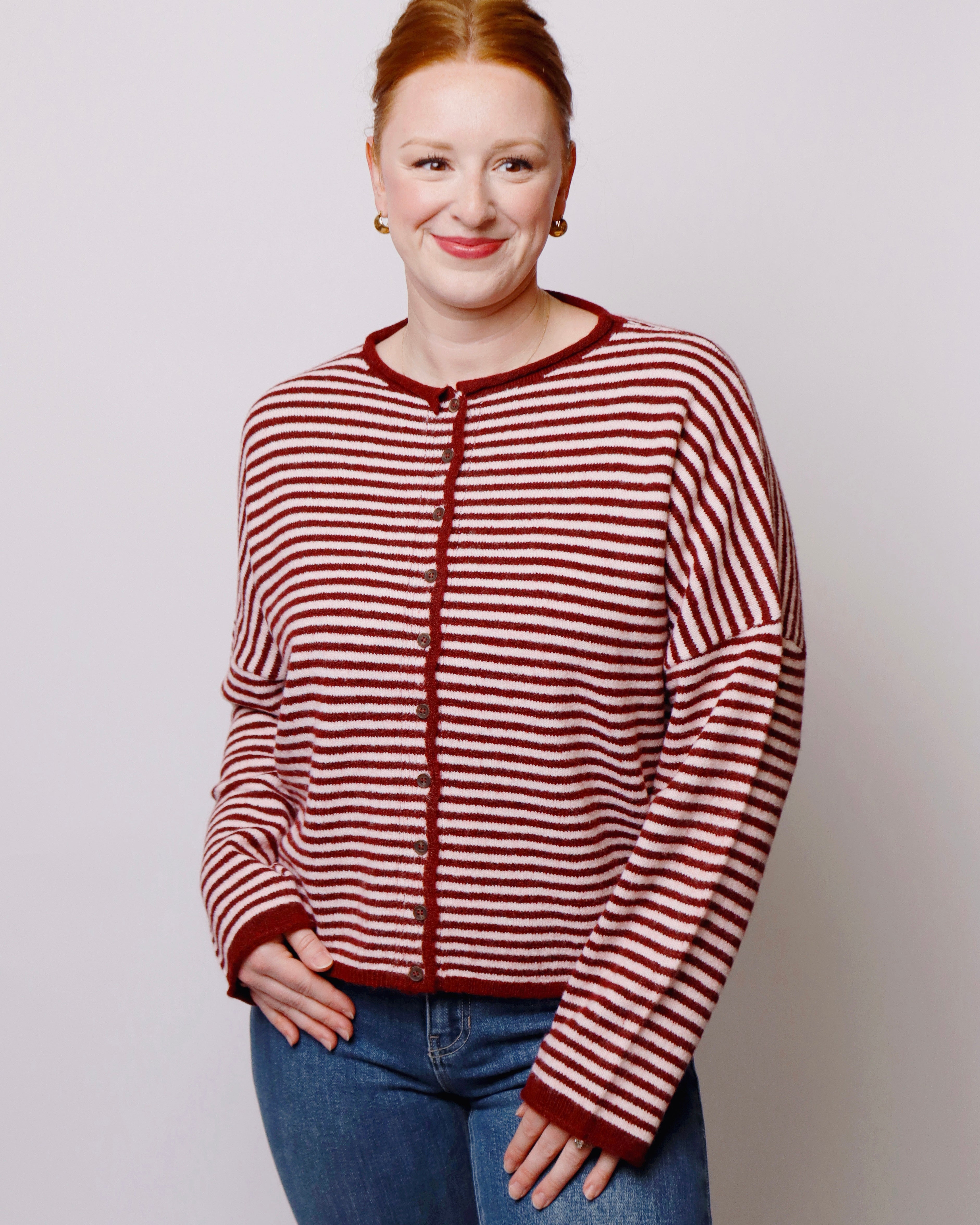 A woman with red hair wearing a red and pink striped knit cardigan sweater and blue jeans, standing against a light gray studio background.