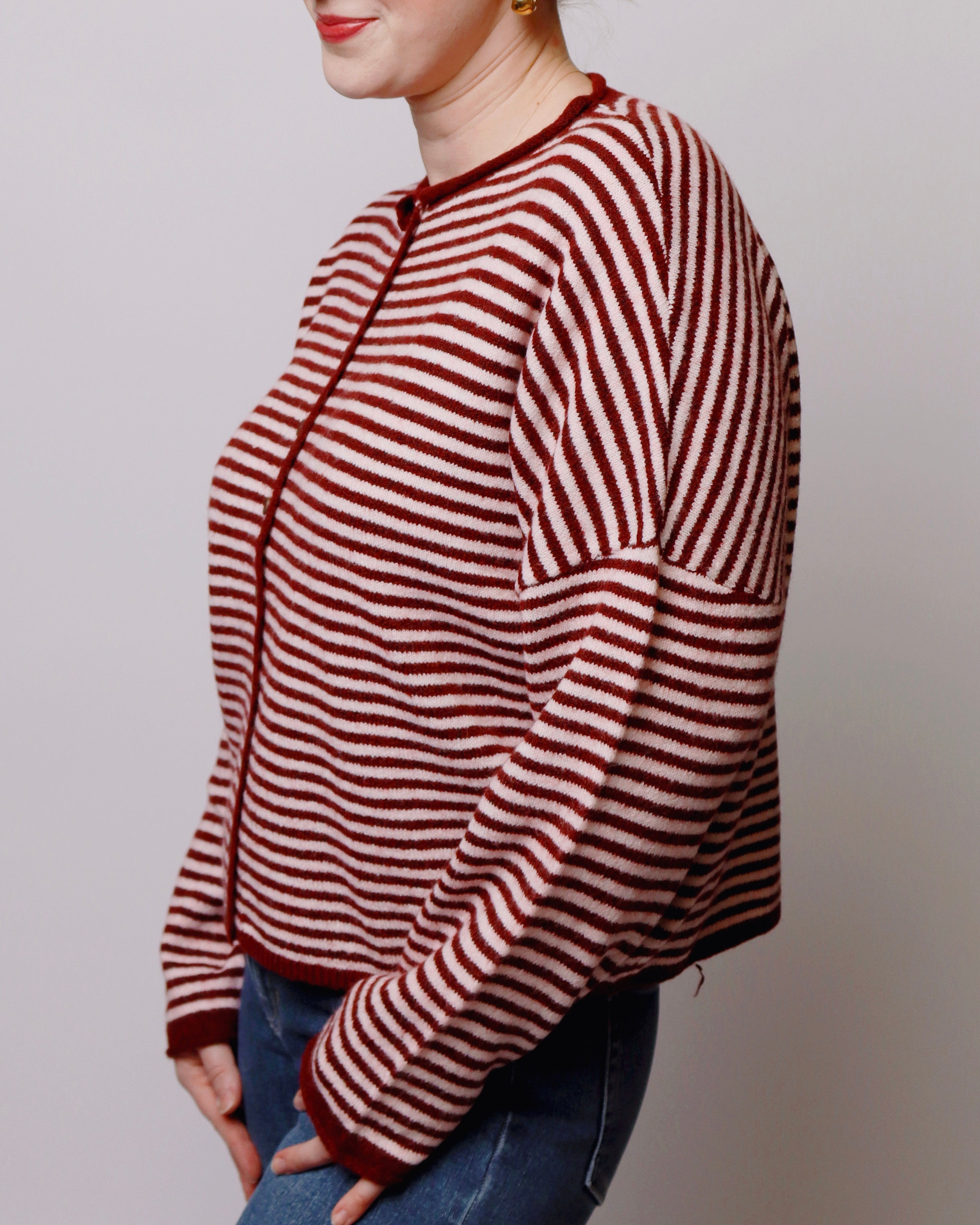 A woman wearing a cropped, long-sleeve knit sweater with horizontal burgundy and pink stripes, paired with blue jeans, shown from a side profile against a plain light gray background.