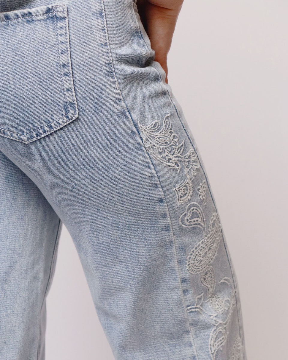Close-up of light-wash blue denim jeans with white embroidered paisley detailing running down the side of the leg.