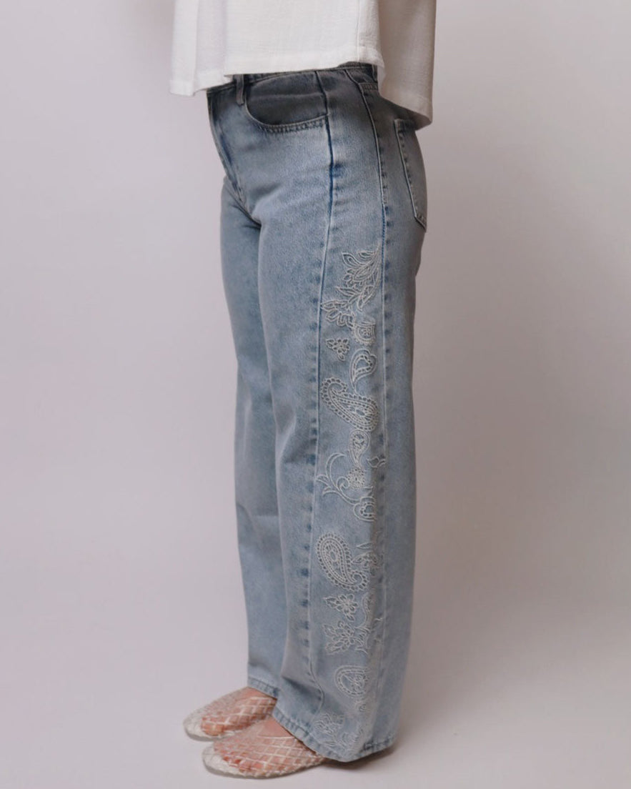 Light-wash blue jeans with ornate white embroidered detailing down the outer side of the legs, shown on a model wearing a white top and patterned slip-on shoes against a plain light gray studio background.