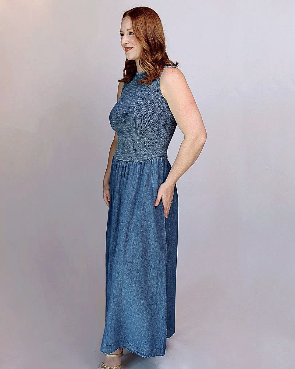 A woman modeling a sleeveless, high-neck, floor-length blue dress with a textured bodice and a flowing skirt, standing against a plain light gray studio background.