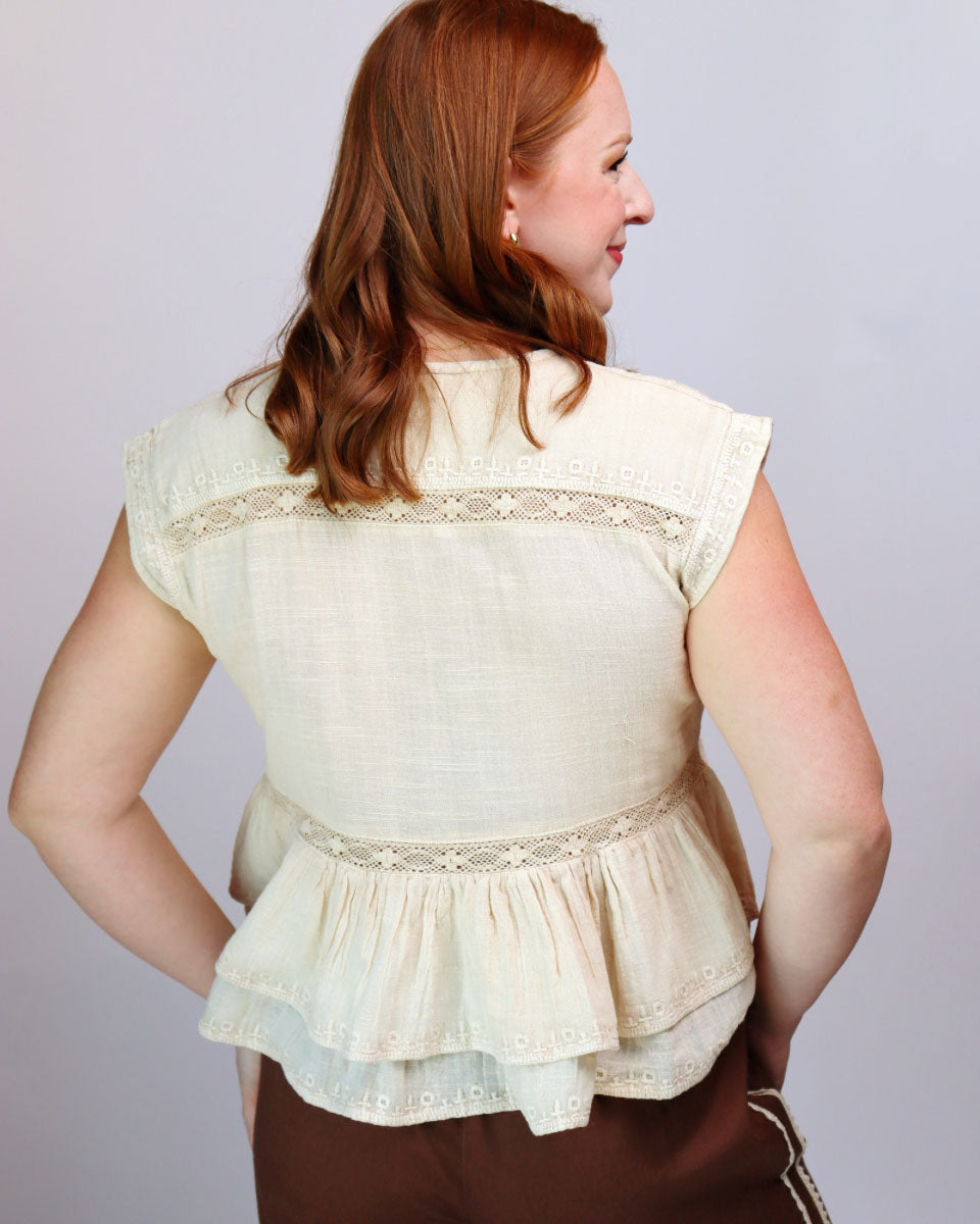 A woman shown from the back wearing a cream-colored peplum blouse with lace trim and ruffled layers, paired with brown pants, against a light gray studio background.