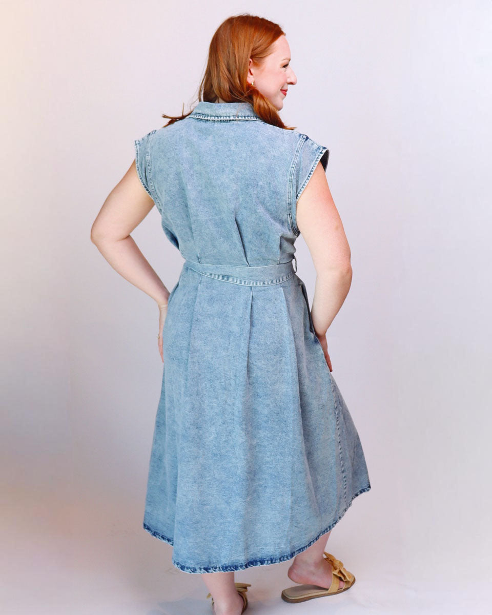 A woman with red hair is shown from the back wearing a light blue denim sleeveless dress with a collar and a matching belt, standing against a plain light background.