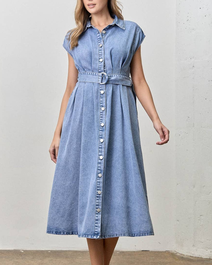 A woman wearing a light blue denim midi dress with a button-down front, short cap sleeves, and a matching fabric tie belt at the waist, standing against a light gray textured wall.