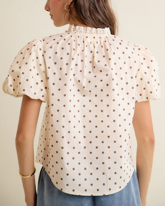A woman shown from the back wearing a cream-colored short-sleeve blouse with brown polka dots and a ruffled high neck, paired with blue jeans against a plain beige background.