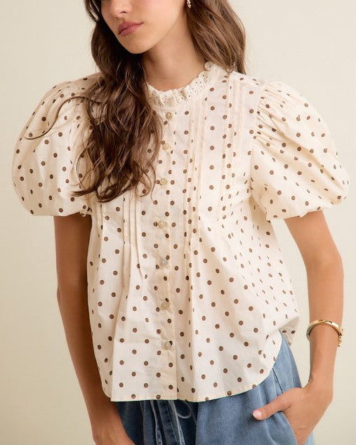 A woman wearing a cream-colored short-sleeve blouse with brown polka dots and puffed sleeves, paired with blue jeans, against a plain beige background.