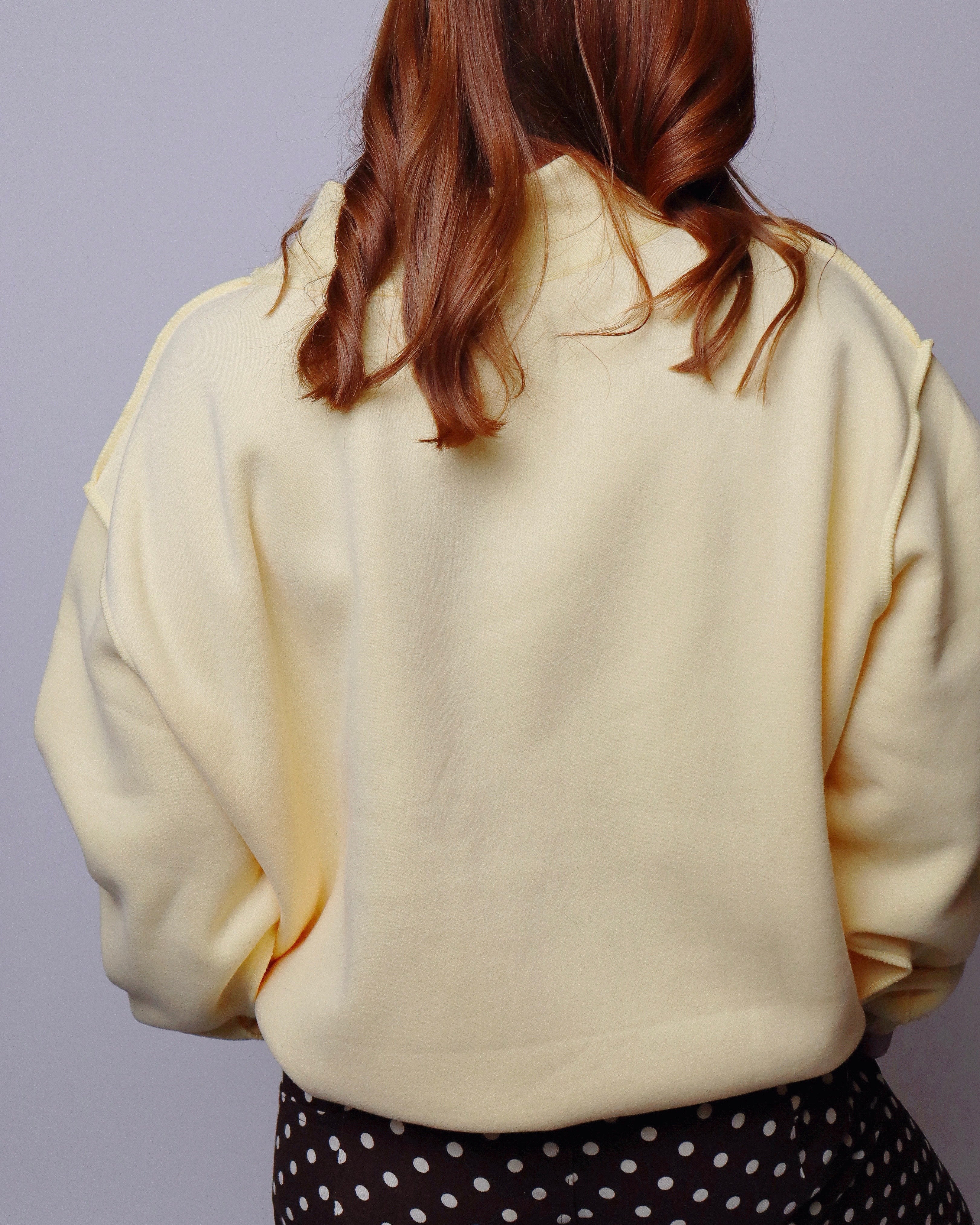 A person shown from the back wearing a pale cream or light yellow oversized sweatshirt, paired with a black and white polka dot skirt, against a solid light gray background.