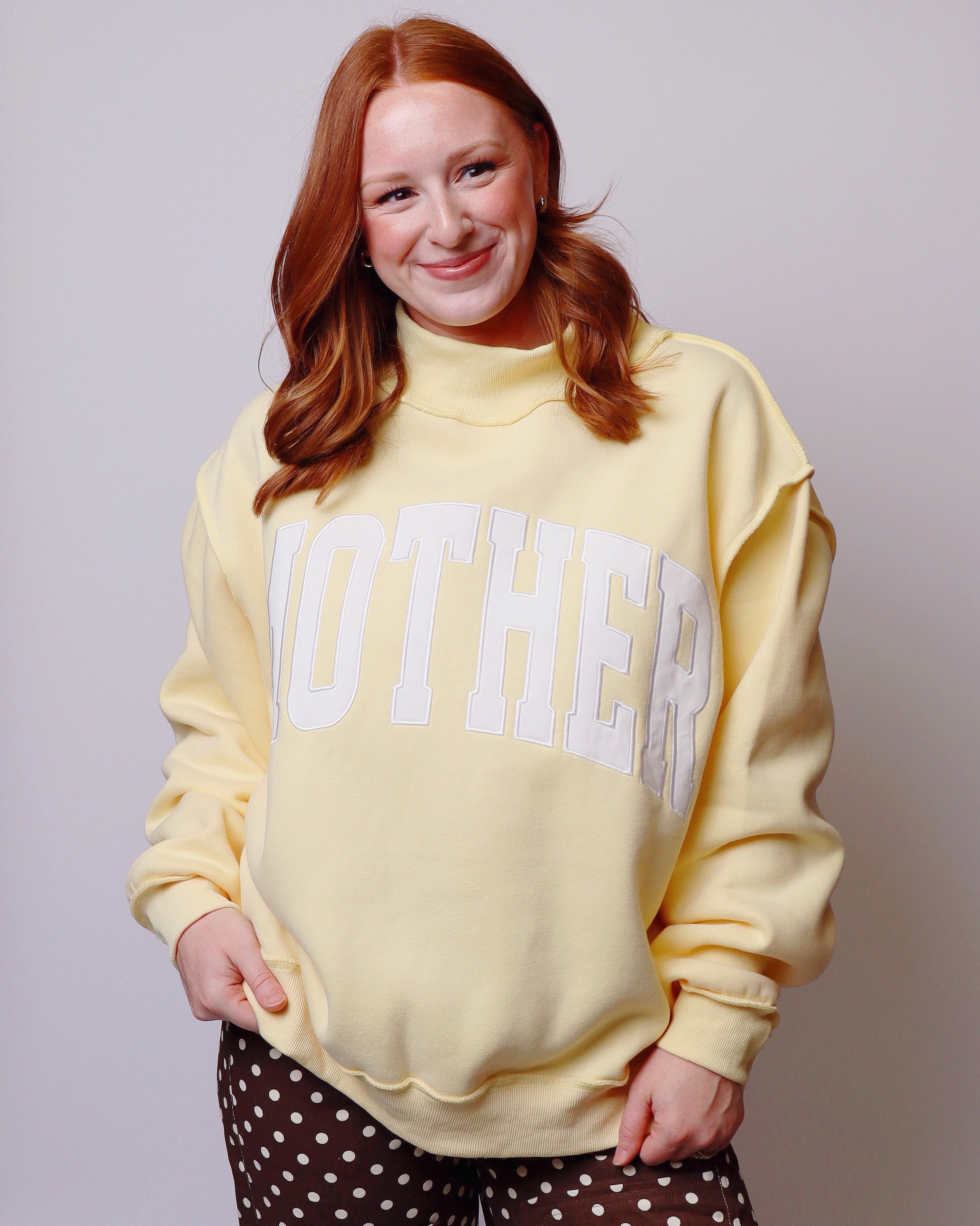 A smiling woman with red hair wearing a pale yellow sweatshirt with 'MOTHER' embroidered in white arched letters across the chest, paired with brown and white polka-dot pants, against a plain light gray background.