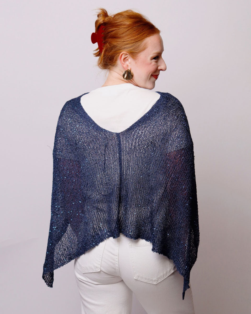 A woman with red hair tied up is shown from the back wearing a sheer, open-knit navy blue shawl over a white top and white pants, against a plain light background.