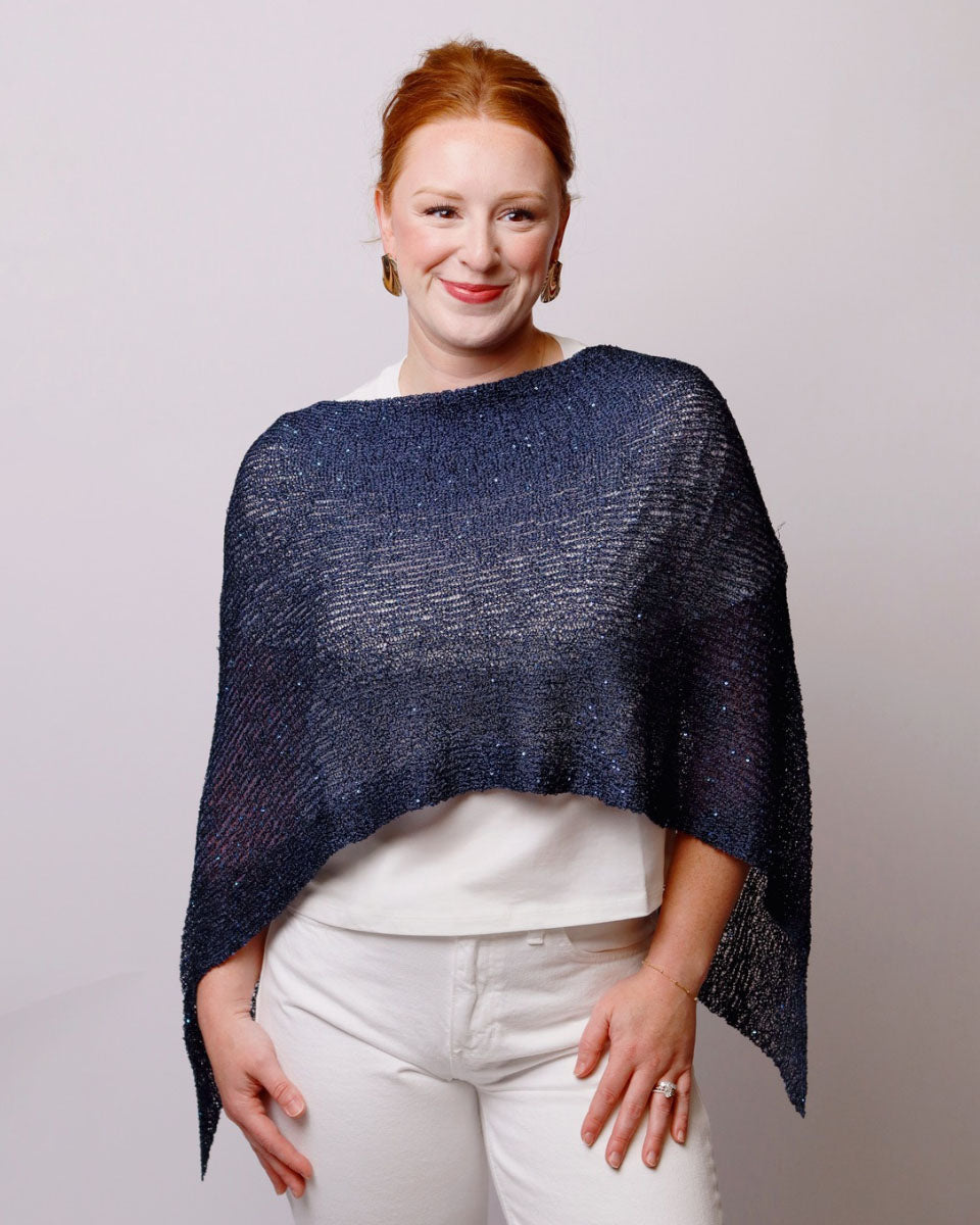 A woman with red hair wearing a shimmering navy blue knit poncho over a white top and white pants, standing against a light gray studio background.