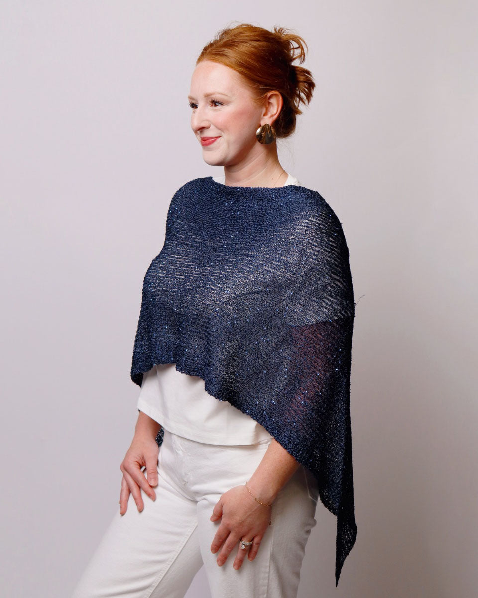 A woman with red hair tied up is wearing a shimmering navy blue knit poncho over a white top and white pants, standing against a light gray studio background.