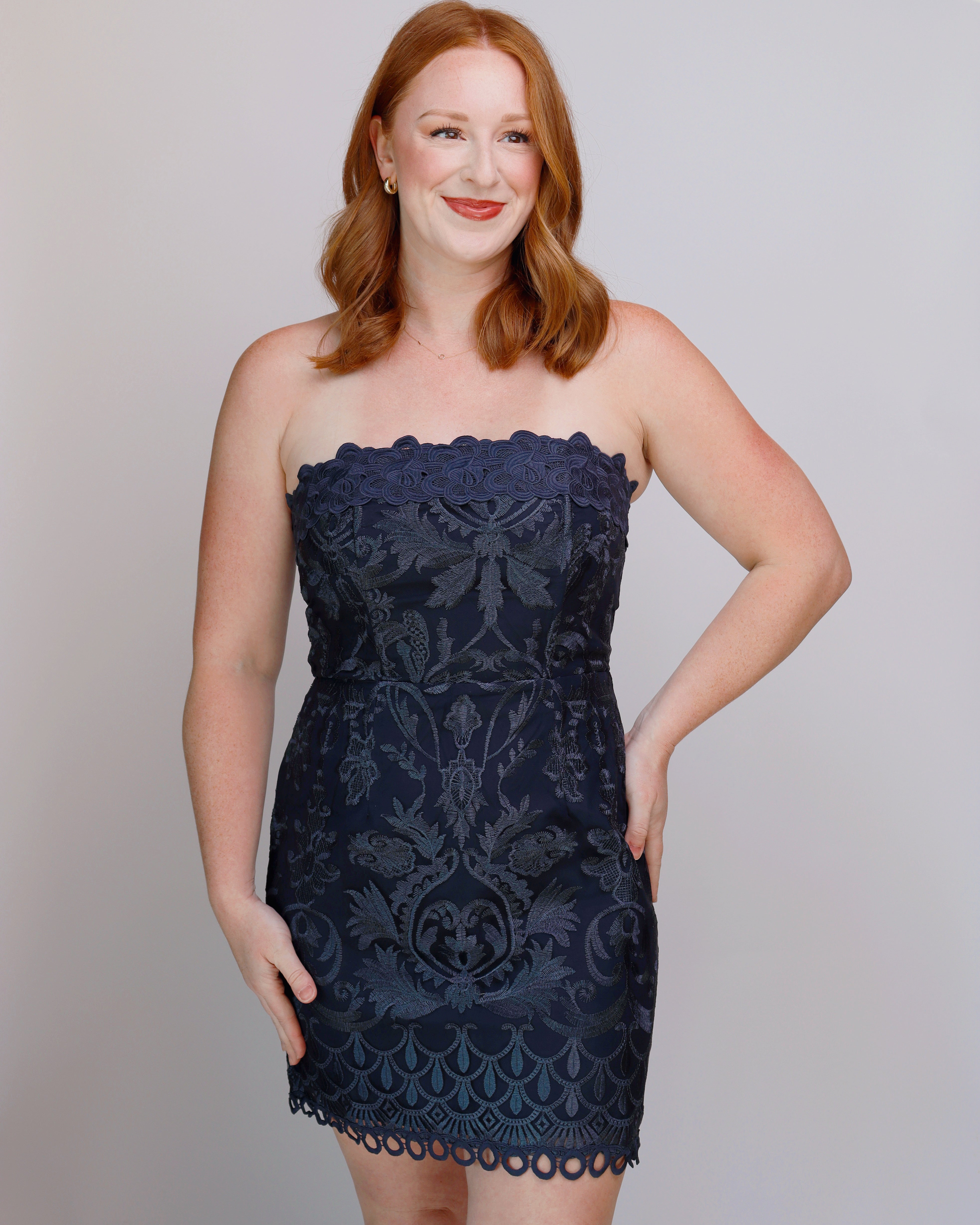 A woman with red hair wearing a strapless dark navy blue lace mini dress with a scalloped neckline and hem, posing against a light gray studio background.