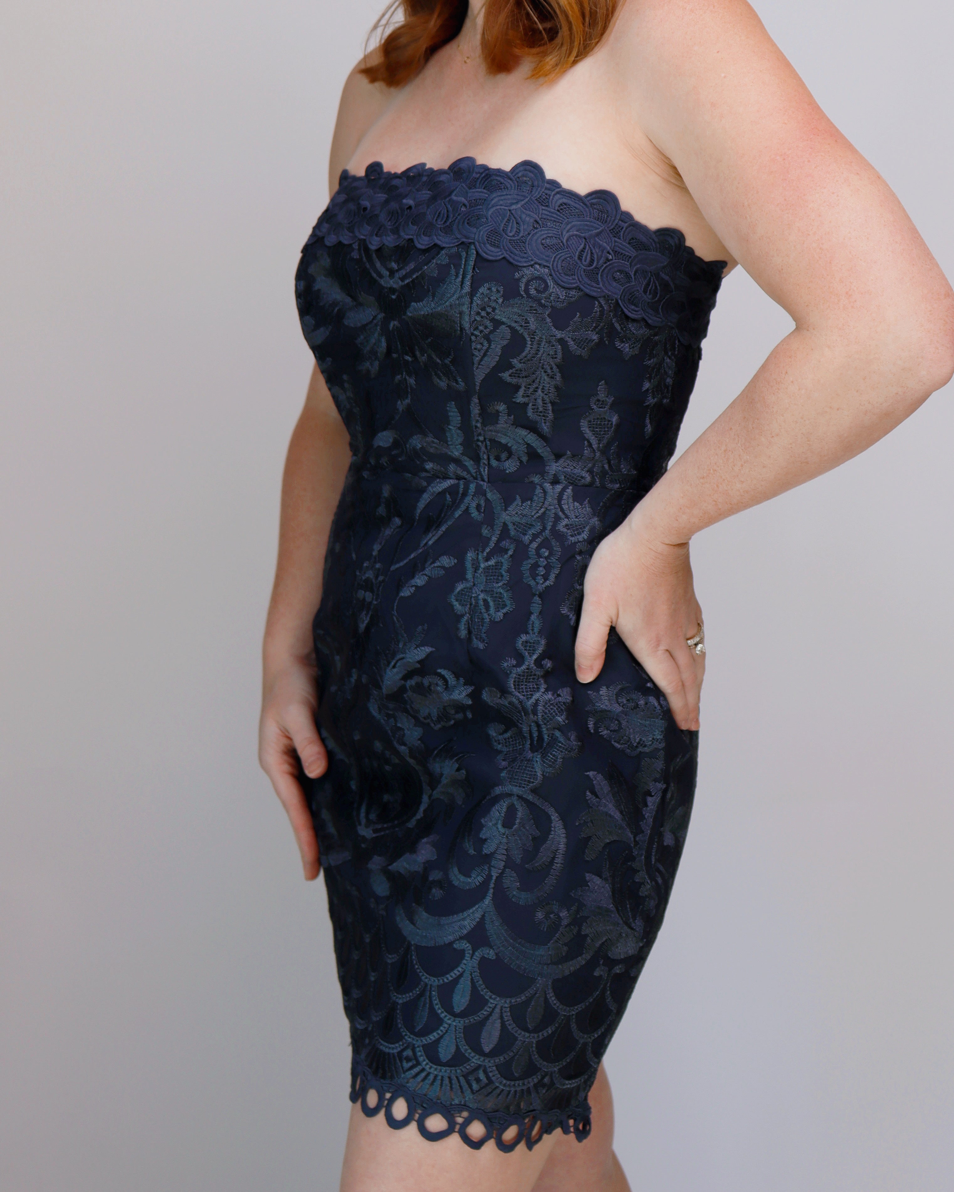 A woman wearing a strapless navy blue lace mini dress with a fitted silhouette and scalloped neckline, photographed against a plain light gray studio background.