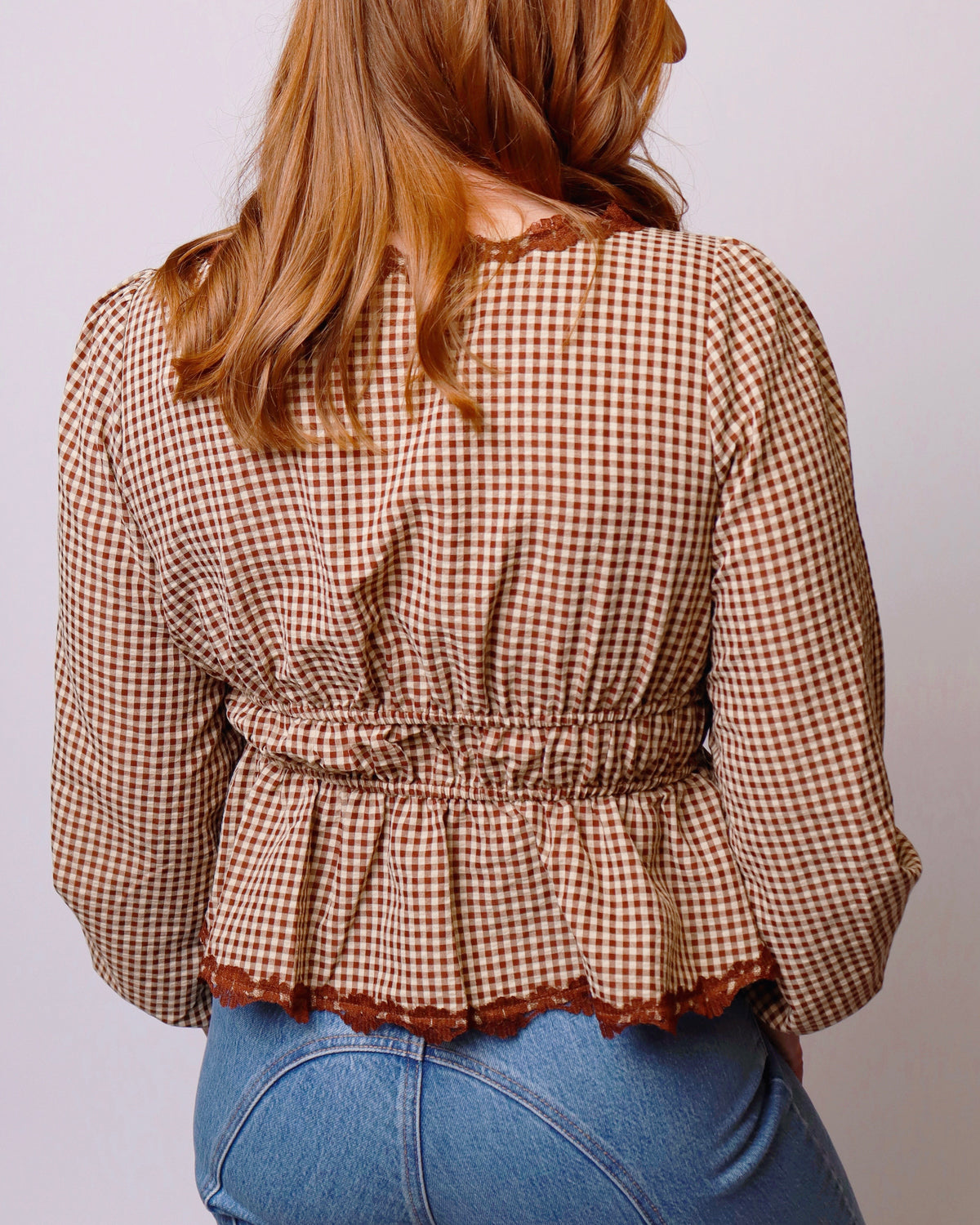 A woman viewed from the back wearing a brown and beige gingham long-sleeve blouse with a smocked waist and ruffled hem, paired with blue jeans.