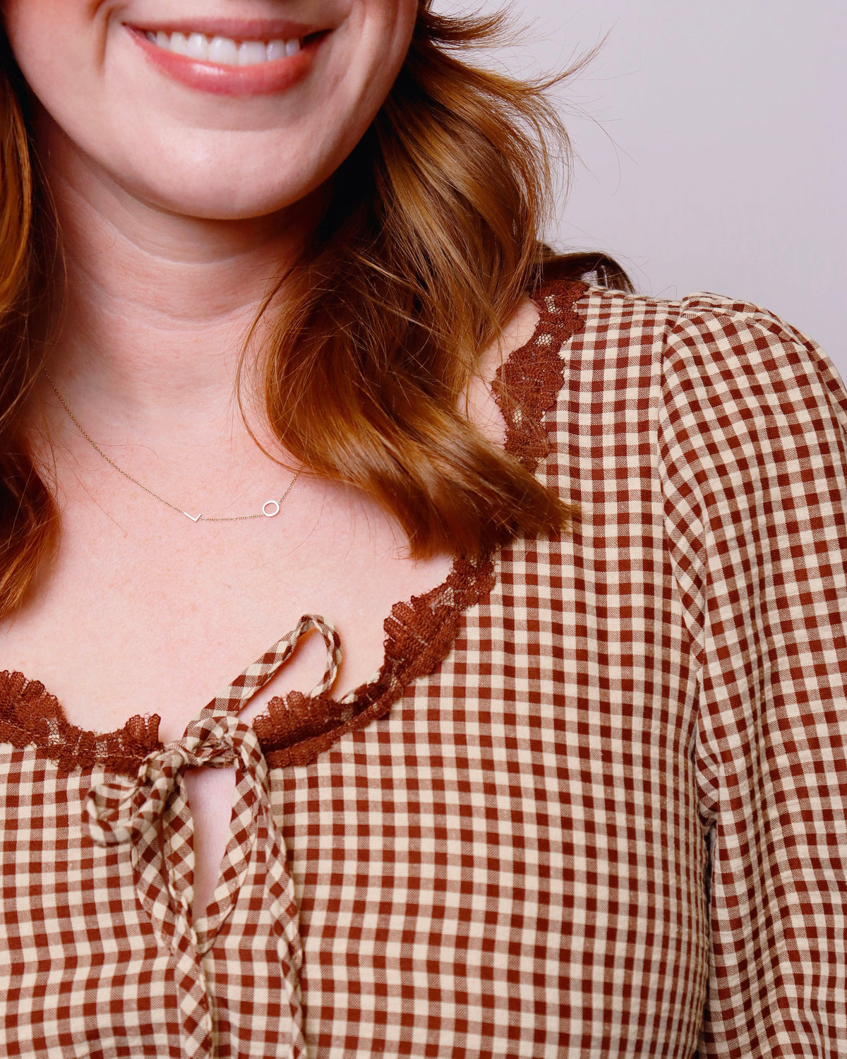 Close-up of a woman wearing a brown and beige gingham top with a tie neckline and brown lace trim, paired with a delicate gold necklace.
